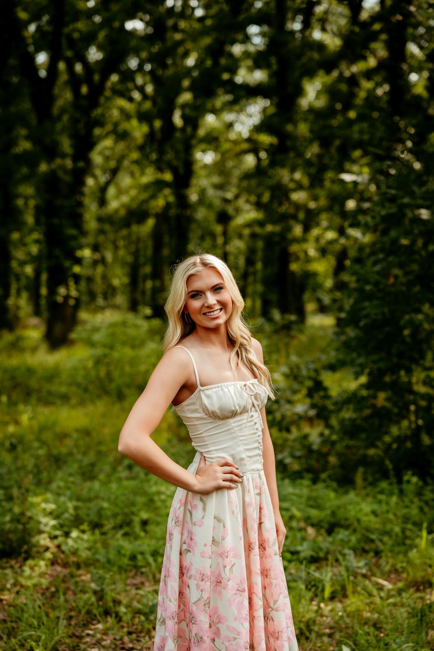 Smiling blonde woman in floral sundress posing in lush green forest setting.