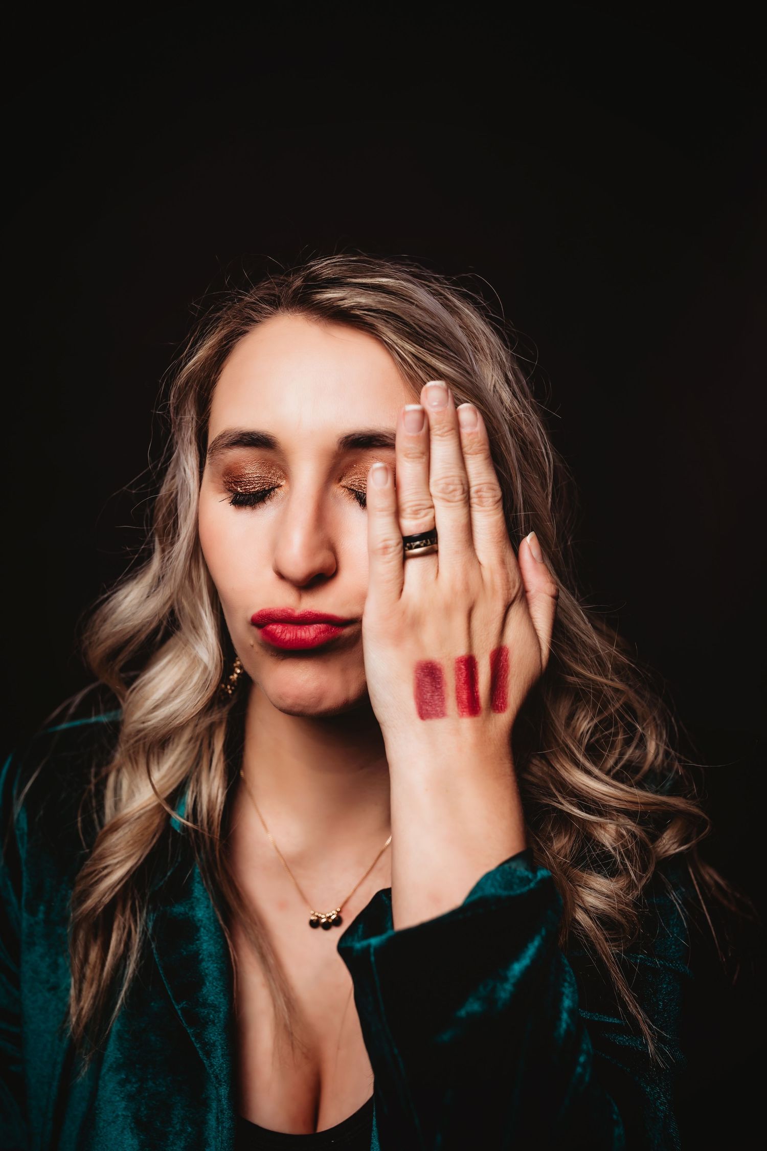 Woman with red lips and lipstick swatches on hand covers half her face, wearing teal velvet jacket.