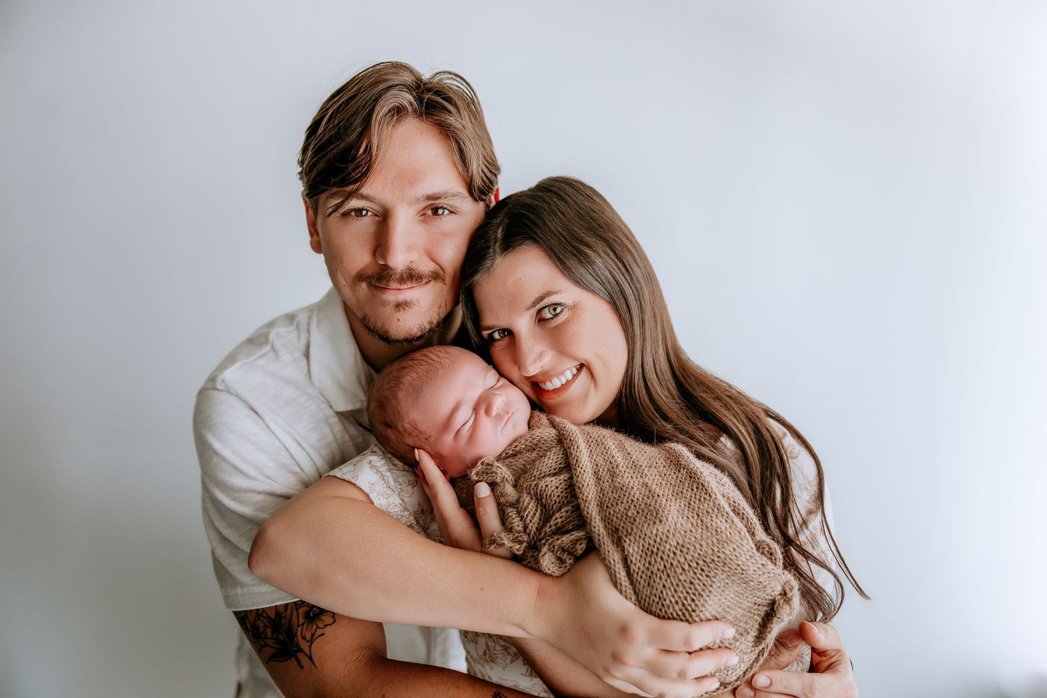 A couple smiles while cradling their sleeping newborn baby, wrapped in a brown knit blanket, against a white background.
