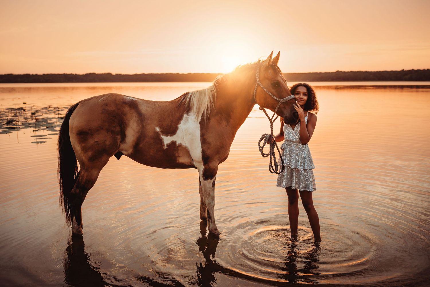 A paint horse stands in shallow water at sunset while someone in a flowy dress pets its neck.
