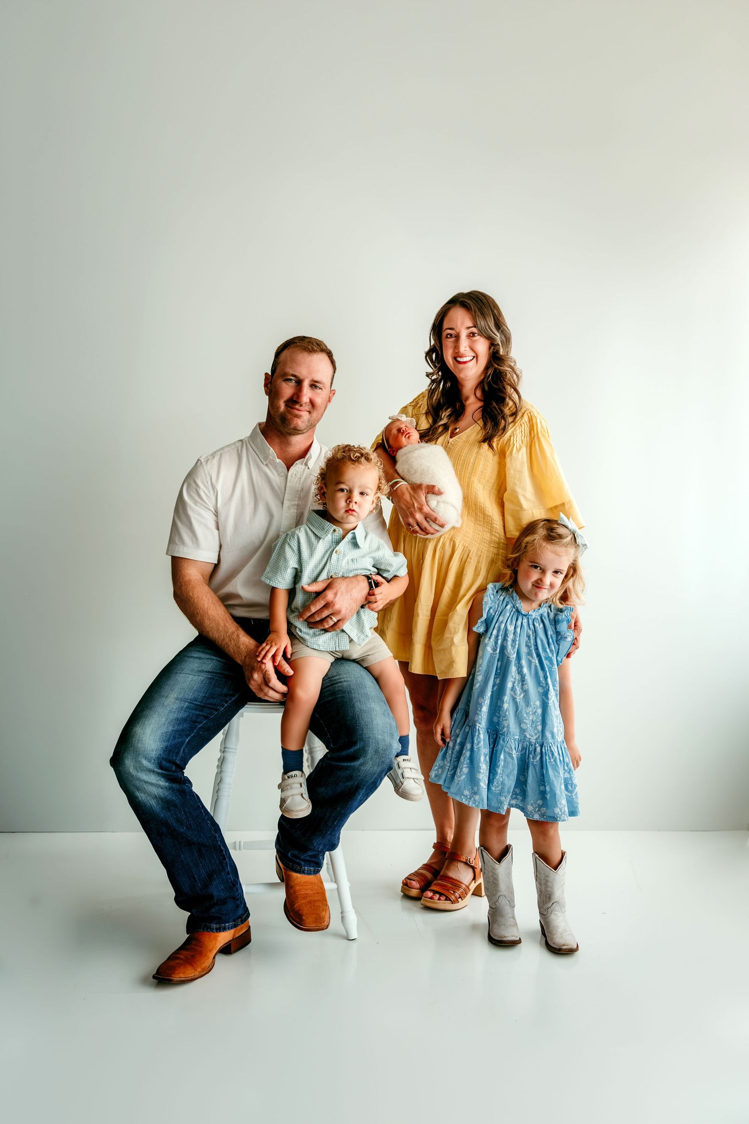 A smiling family of five poses in a bright studio, with parents holding two toddlers and an older girl in a blue dress.