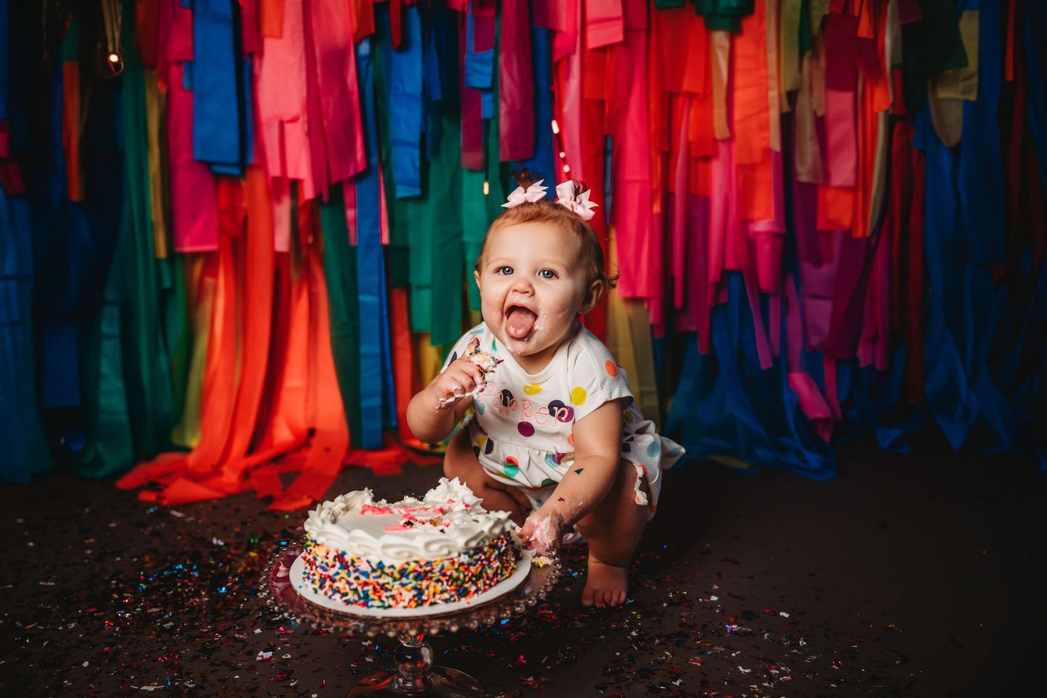Baby girl in polka dot dress smashing first birthday cake with colorful streamer backdrop and sprinkles.
