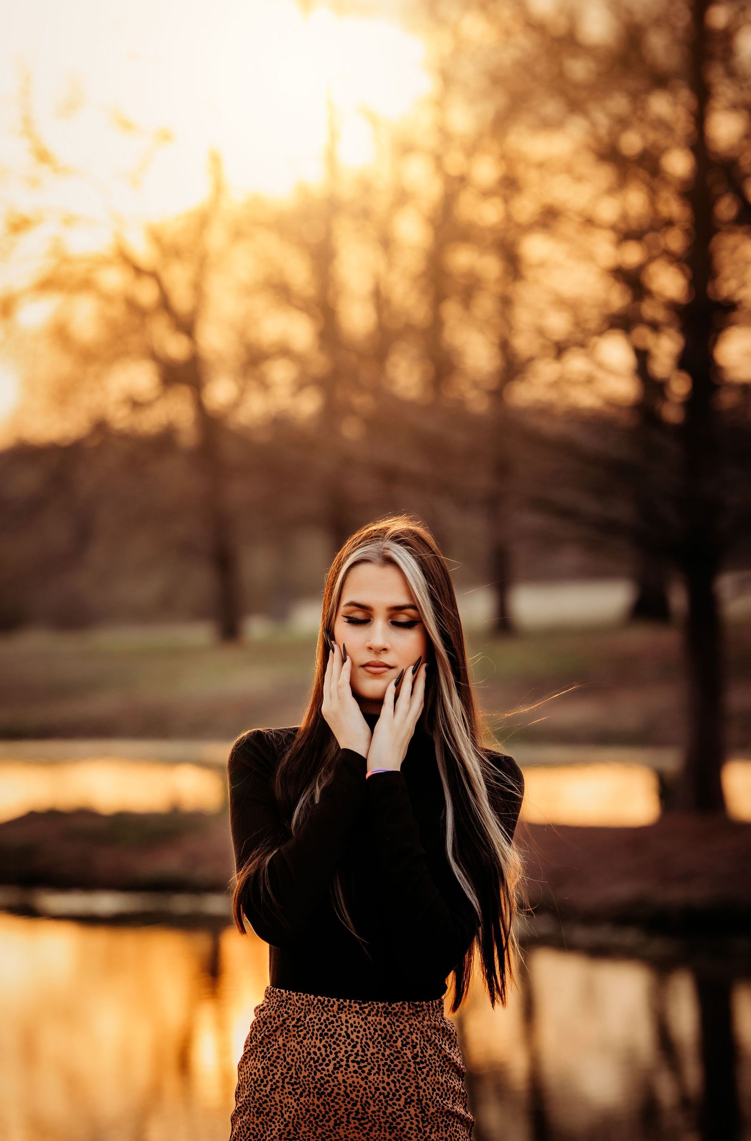 Young woman with long hair poses at golden hour sunset near a reflective pond with bare trees in background.