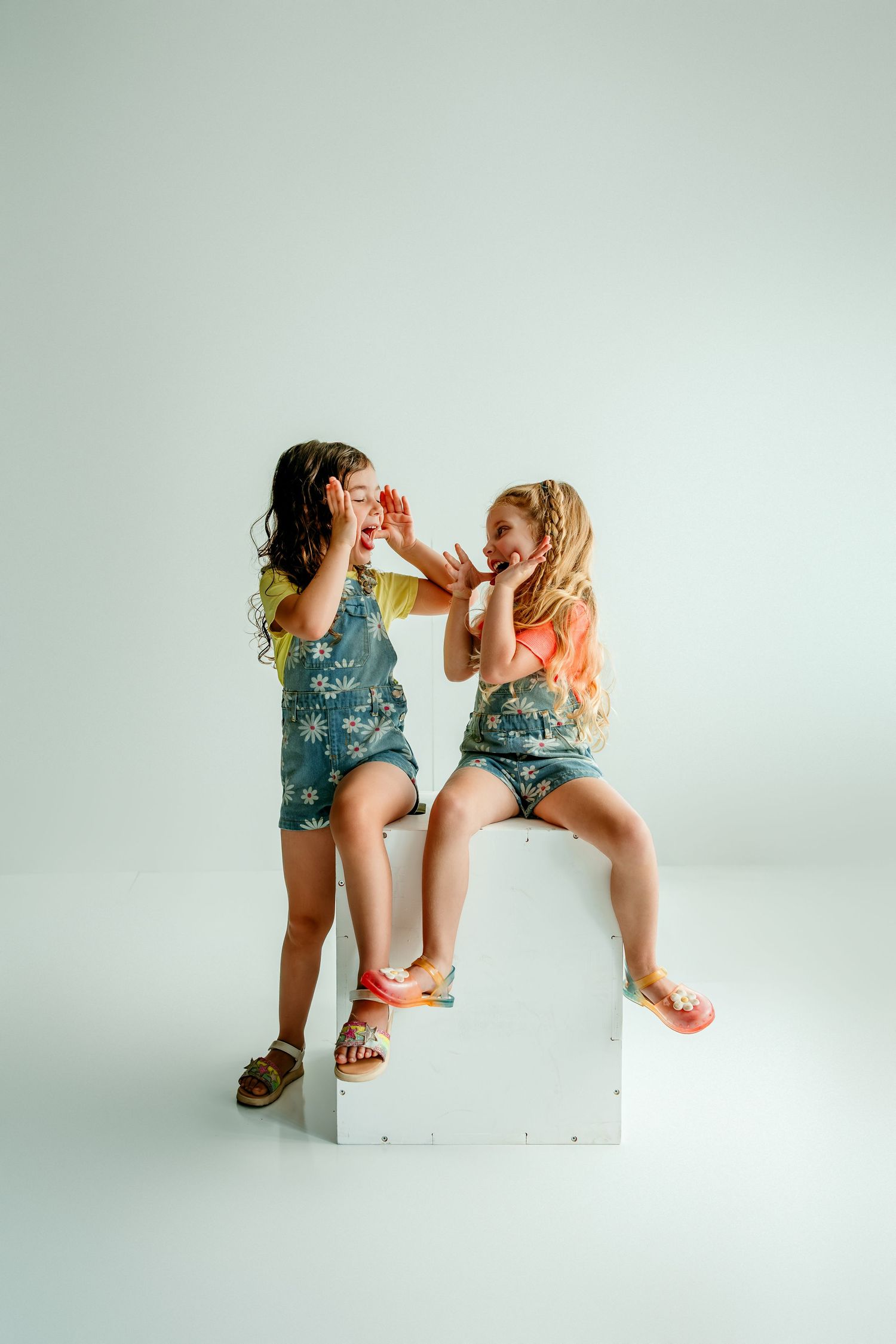 Two young girls in denim floral overalls sit on a white cube, playfully sharing a snack in a studio setting.