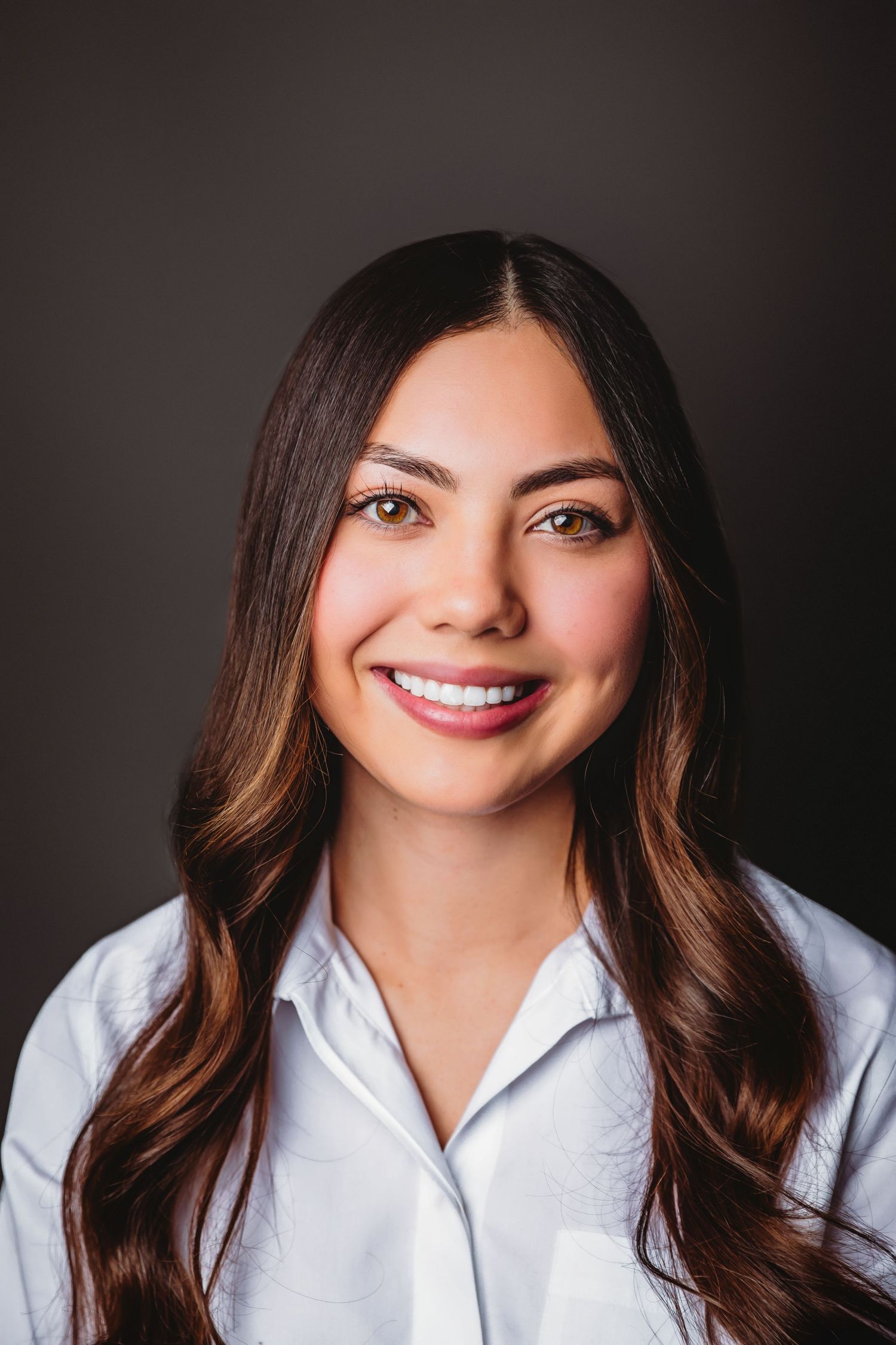 Smiling young woman with long dark hair wearing a white button-up shirt against a dark gray background.