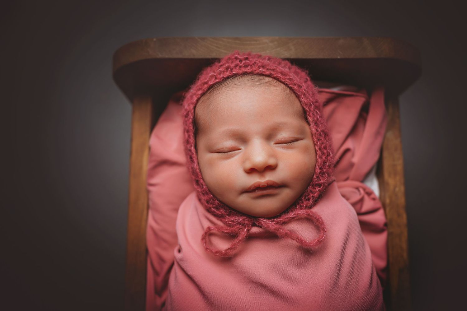 Newborn baby swaddled in pink wrap and bonnet, sleeping peacefully in a wooden box during a studio photo session.