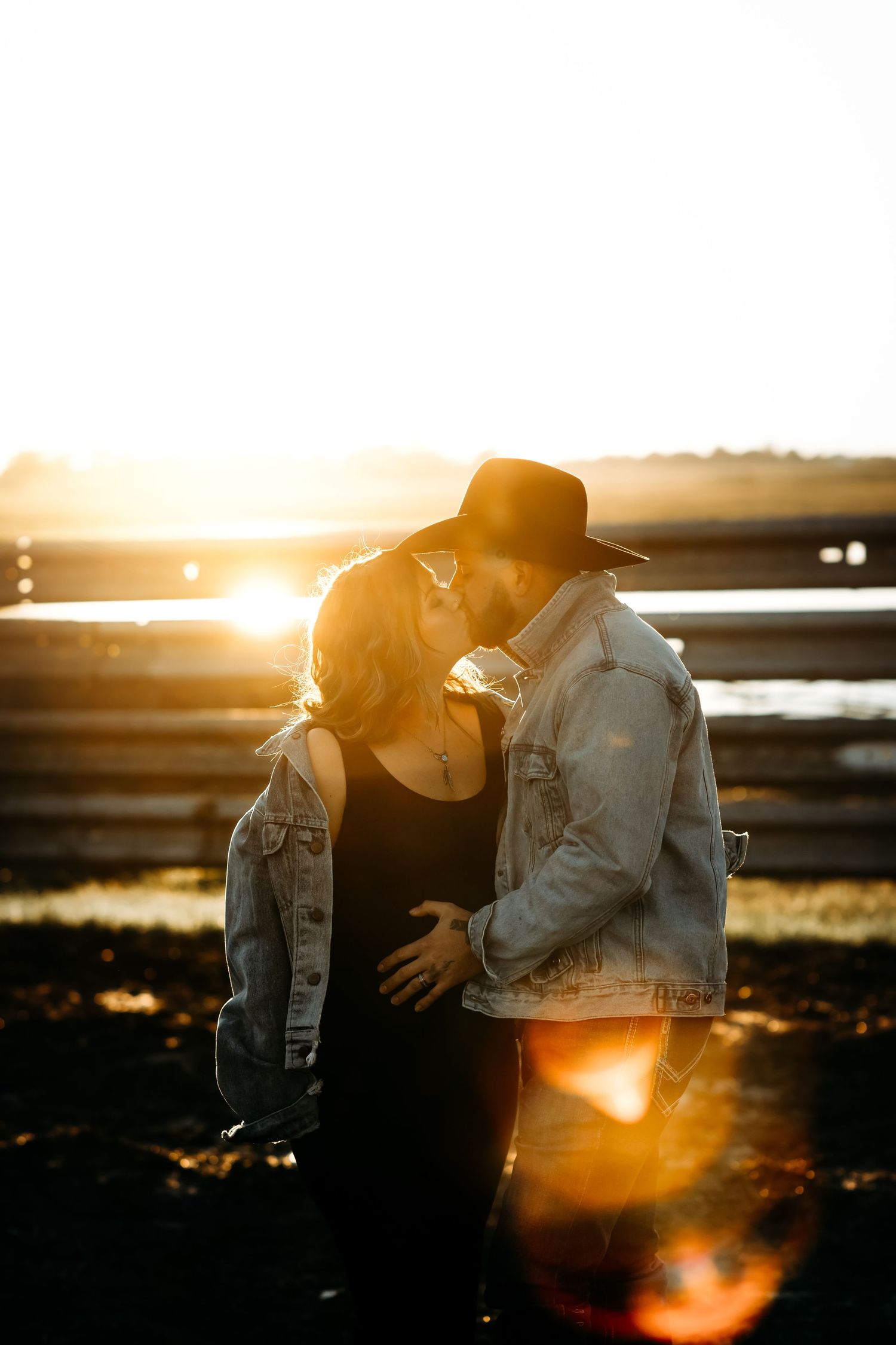 Couple sharing a romantic kiss at golden hour sunset, woman in black dress and man in denim jacket and hat.
