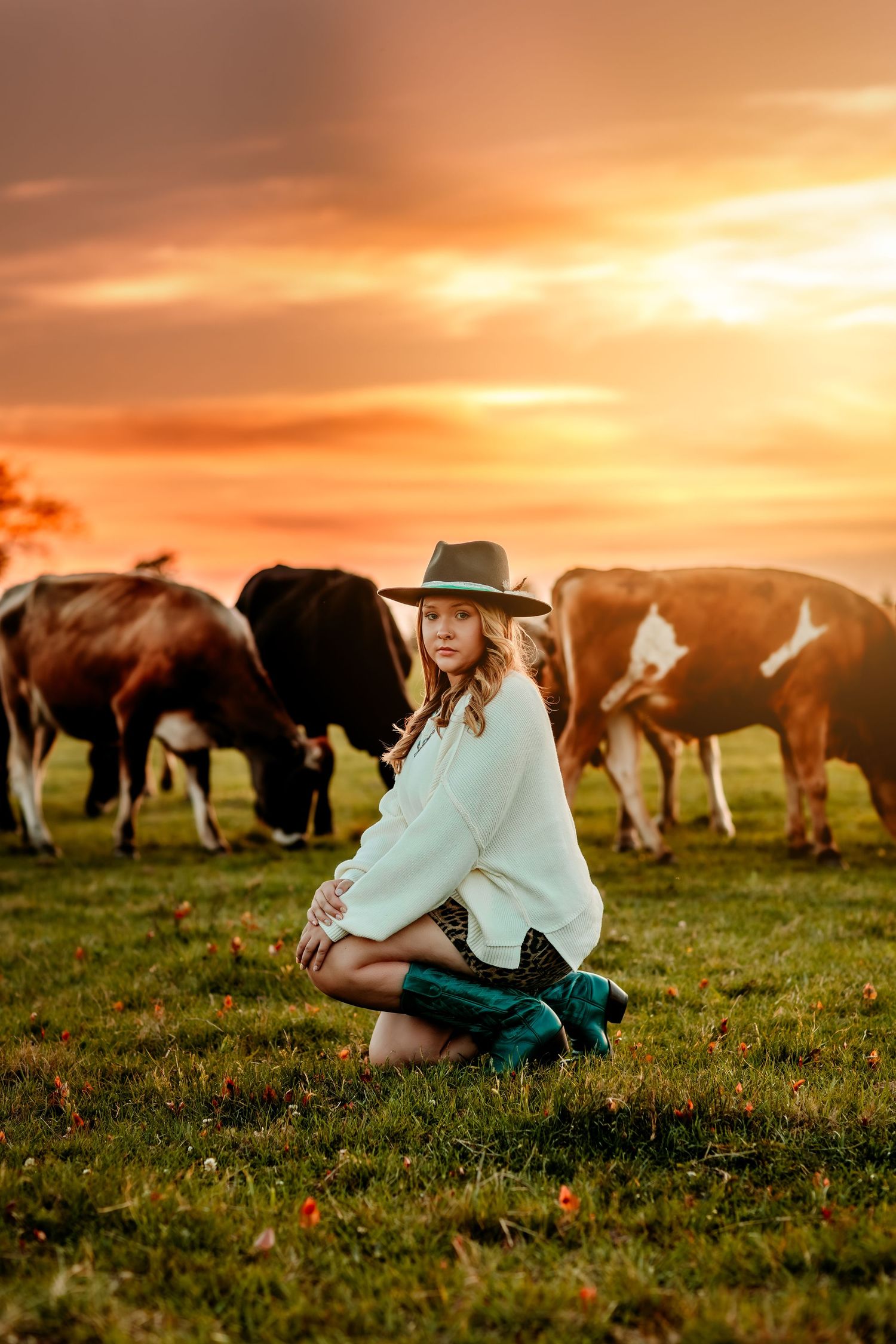 A young woman in a cowboy hat kneels on green grass with grazing cows behind her under a vivid orange sunset sky.