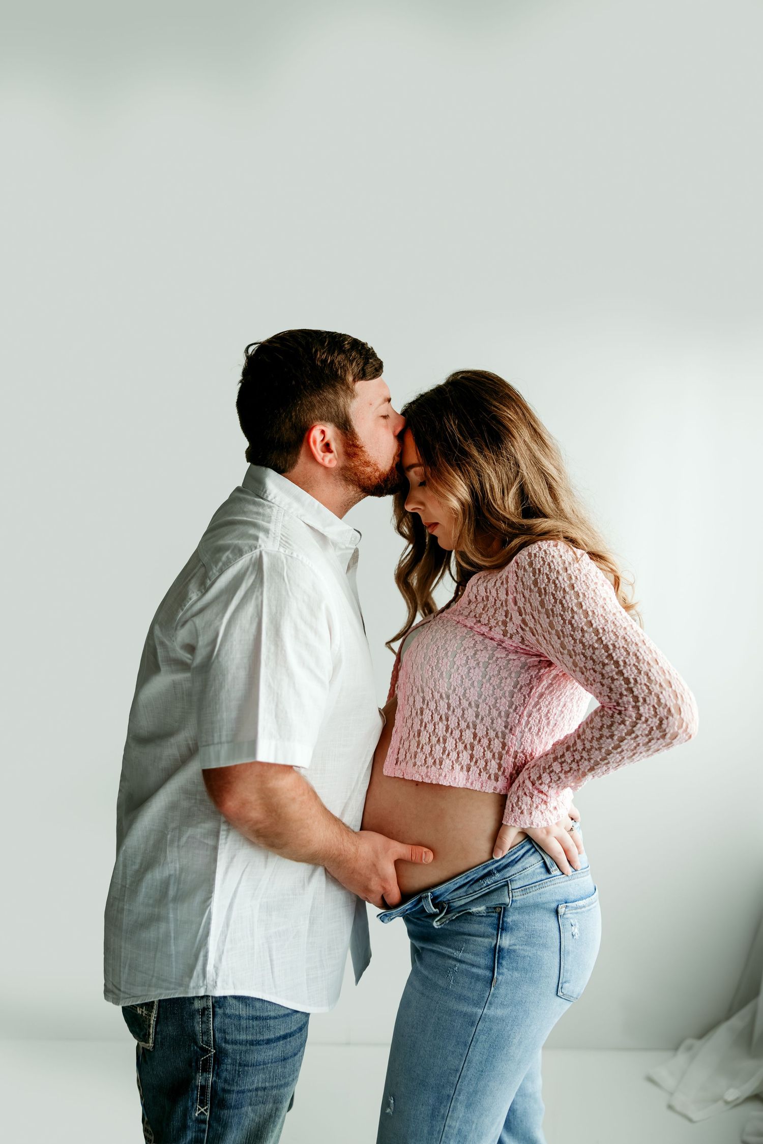 A couple shares a tender kiss while the woman displays her baby bump in a bright studio maternity photo session.