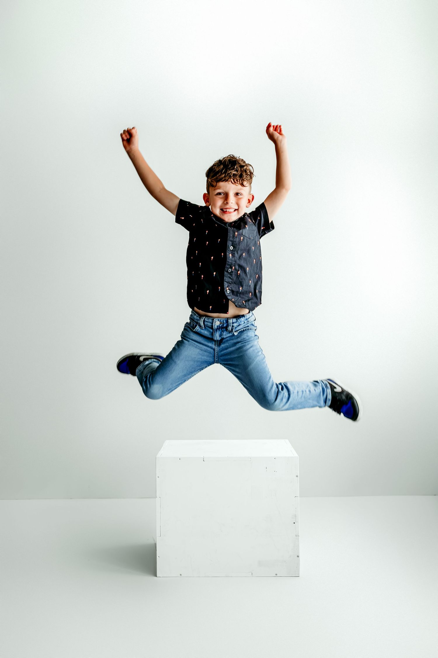 Excited boy jumping off a white cube with arms raised, wearing jeans and black t-shirt, against white background.