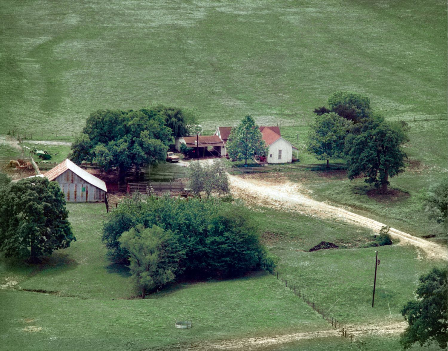 Aerial view of a winding dirt road leading through green rural farmland with scattered buildings and trees.