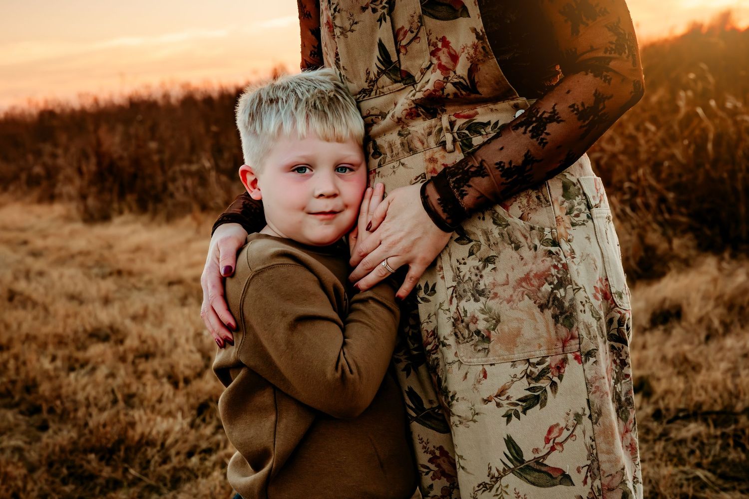 A young child in a brown sweater hugs someone wearing a floral dress during golden hour sunset in a field.