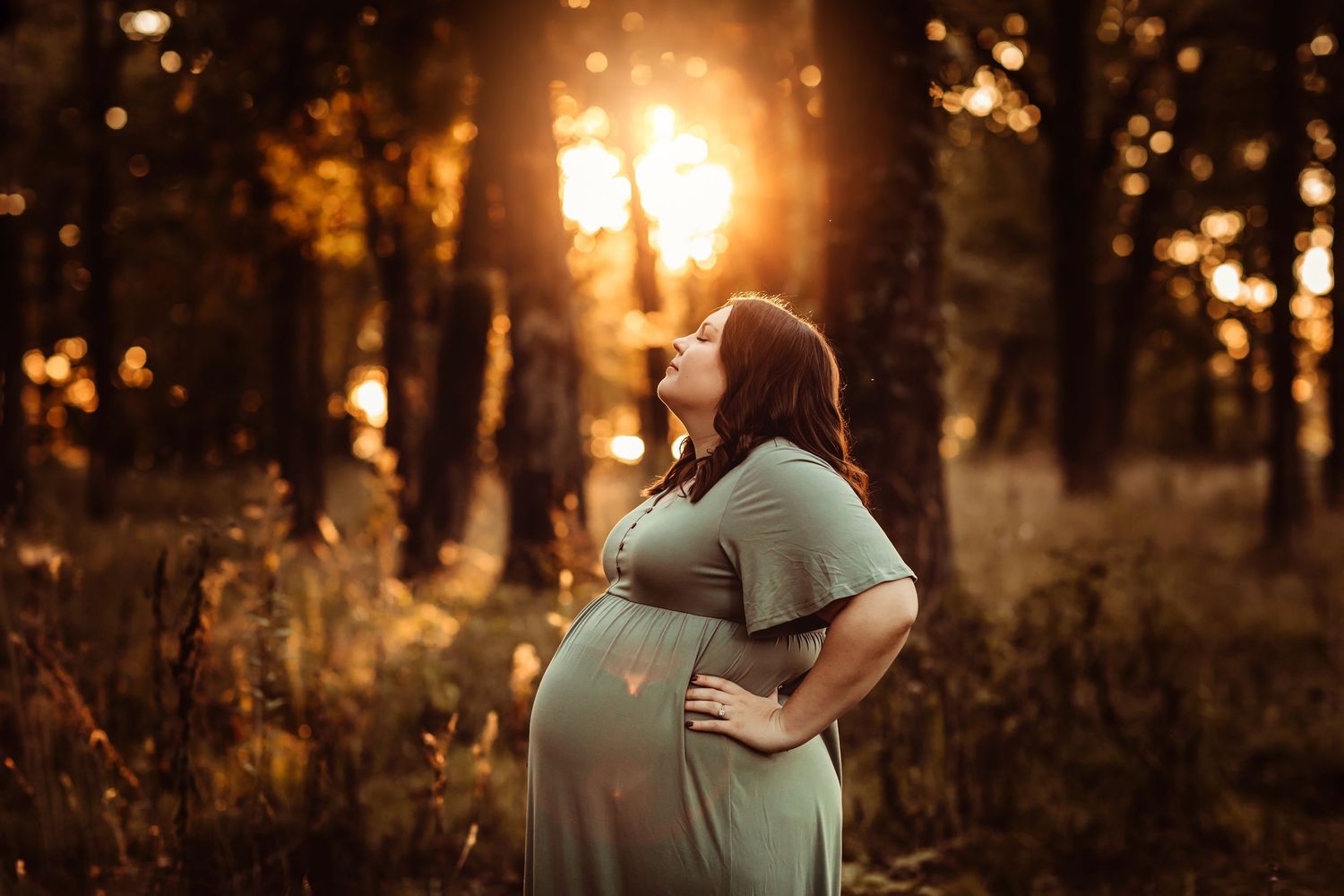 Pregnant woman in green dress standing in sunlit forest at golden hour, hands on hips, looking up peacefully.