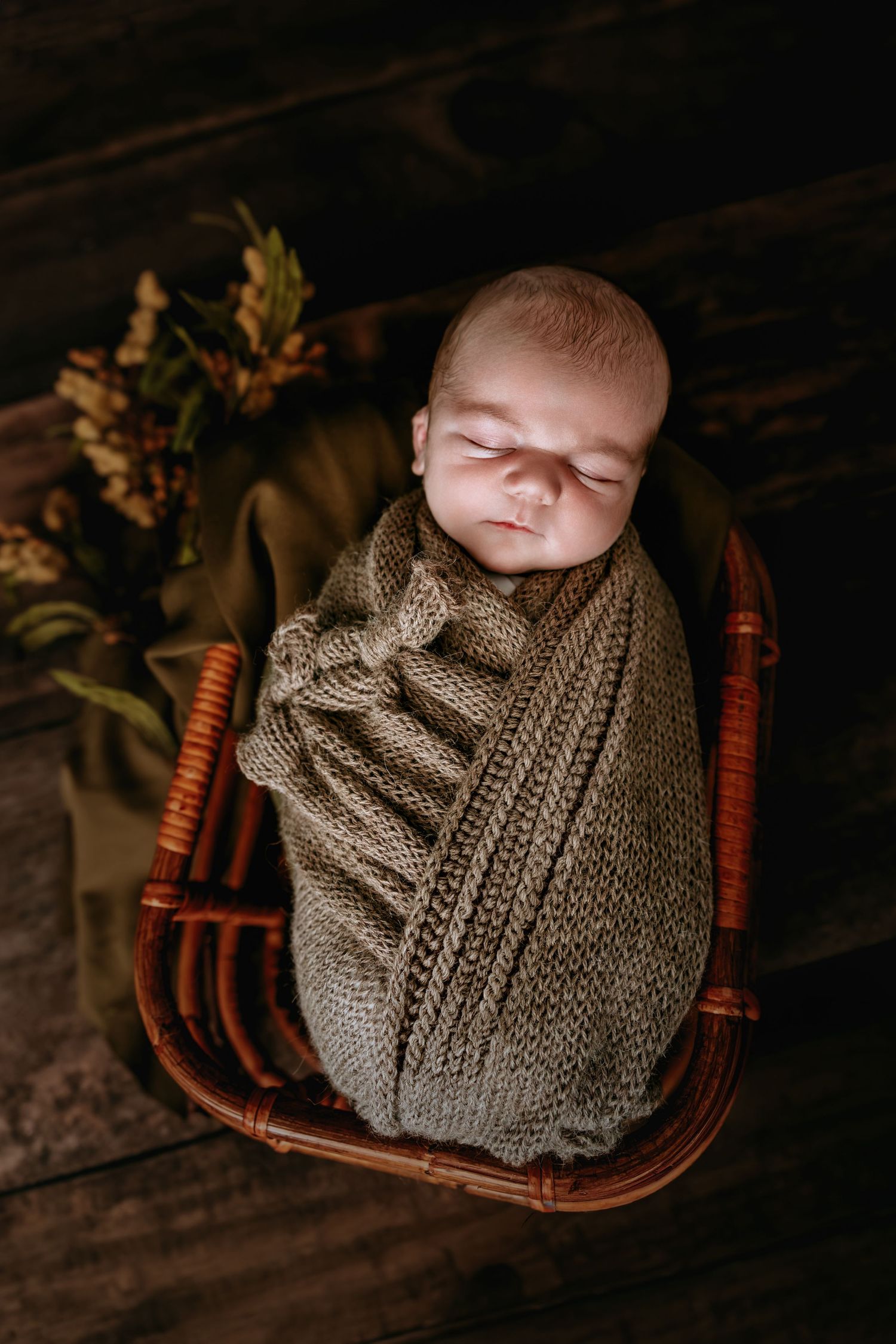 Newborn baby swaddled in olive knit wrap sleeping peacefully in wicker basket with autumn foliage backdrop.