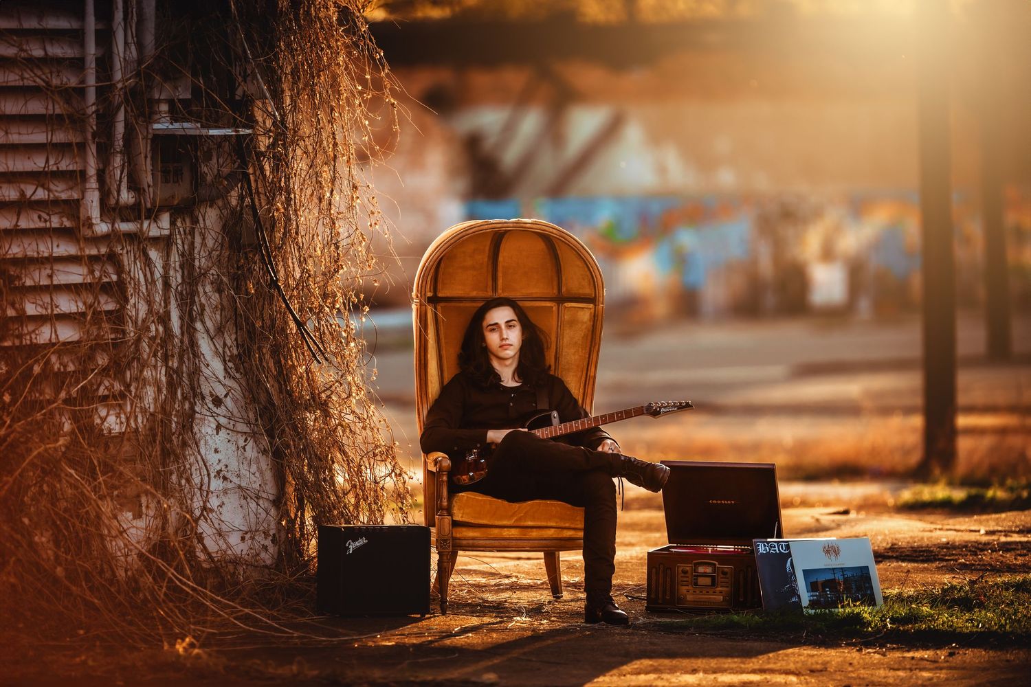 Person with long dark hair sitting in a vintage chair outdoors at golden hour, holding a guitar with luggage nearby.