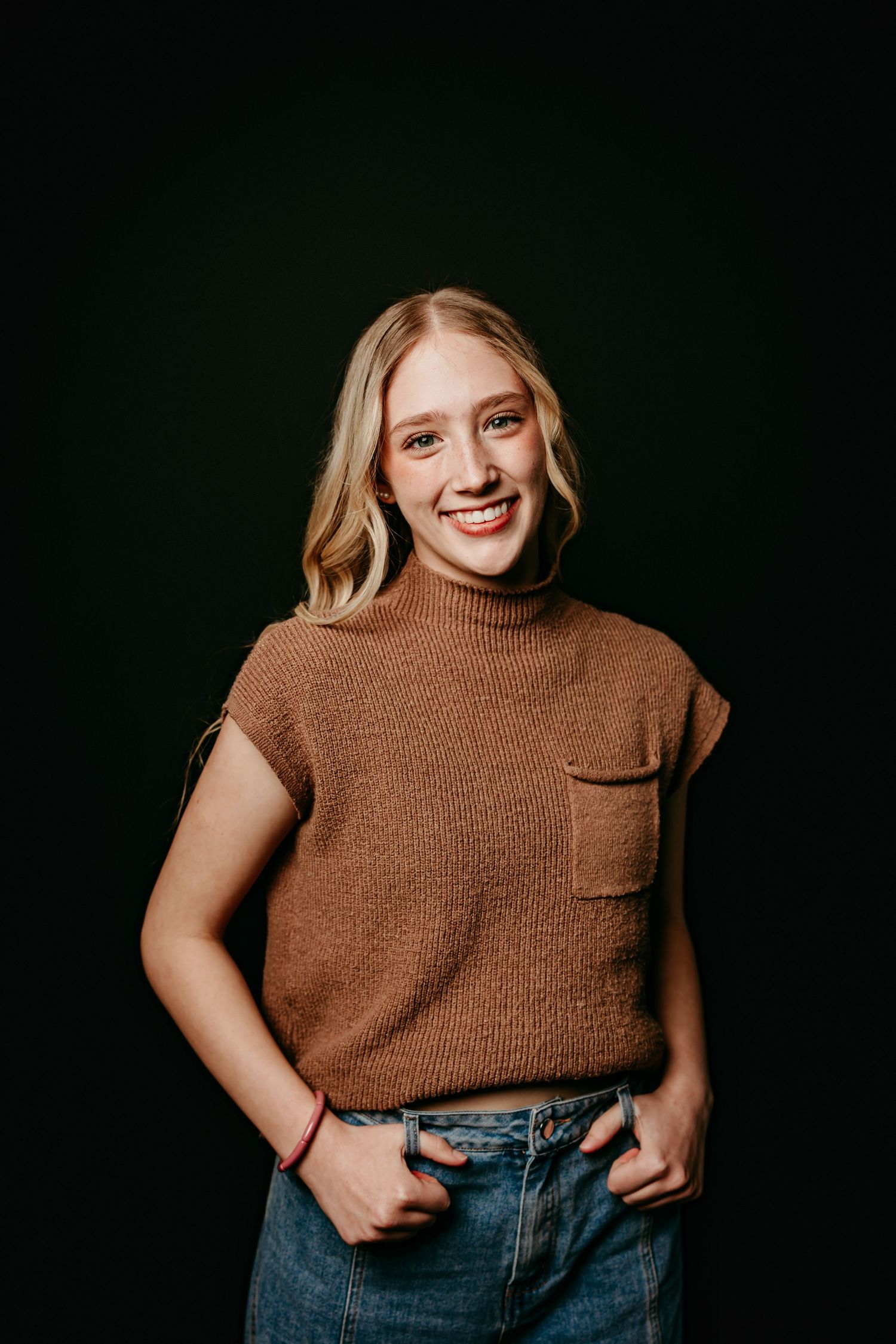 Smiling young blonde woman in brown knit sleeveless sweater and jeans posing against black background.