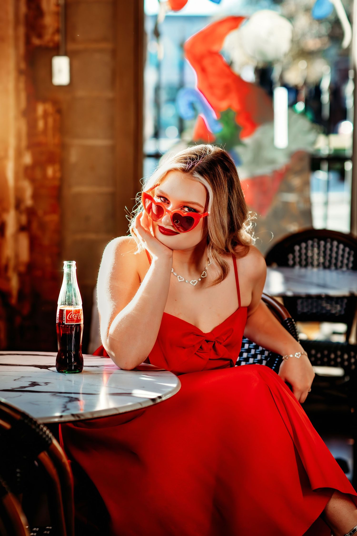 Woman in red dress and heart sunglasses sitting at cafe table with Coca-Cola bottle in sunny outdoor setting.