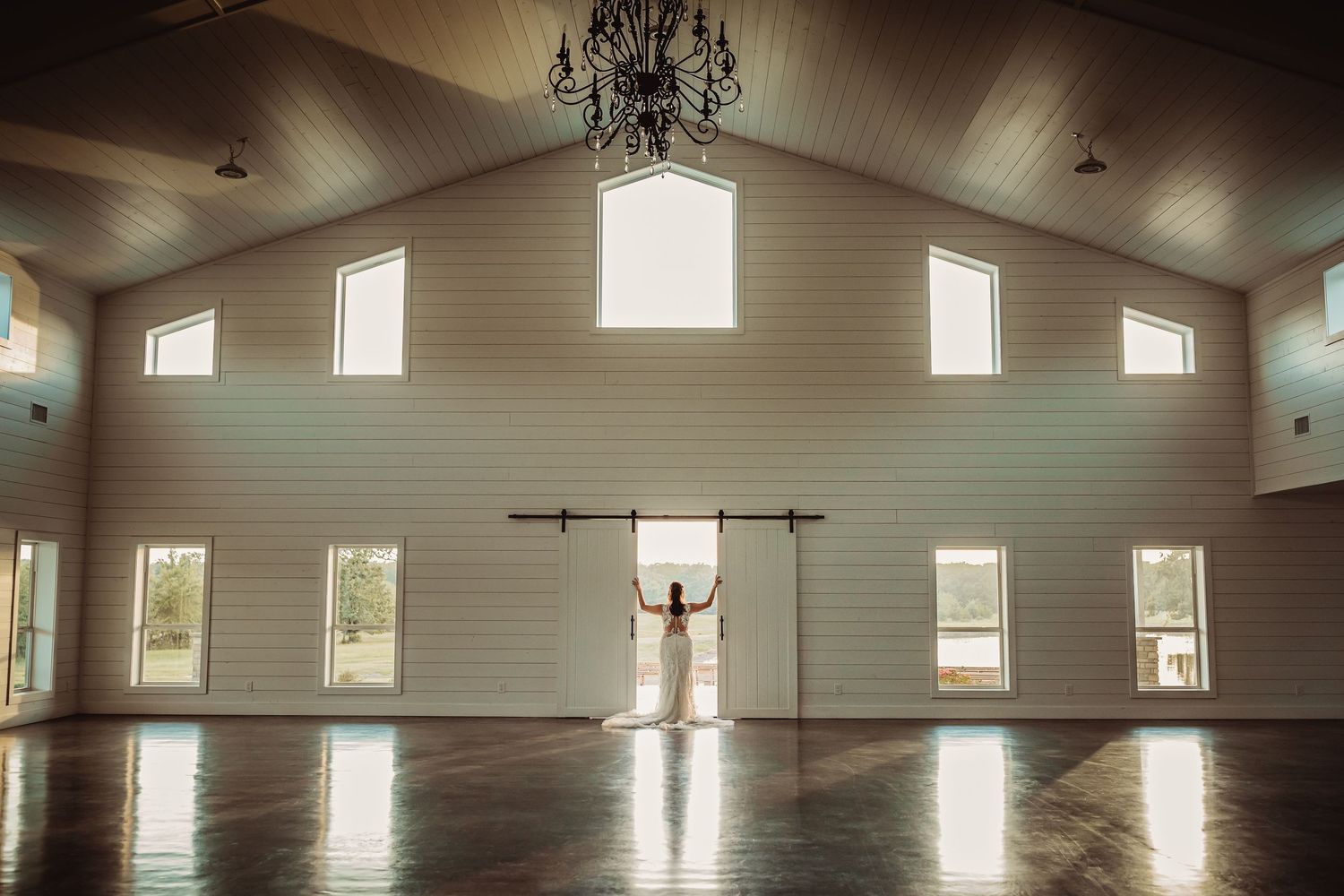 Bride stands in sunlit barn wedding venue with vaulted ceilings, chandelier, and open barn doors creating dramatic silhouette.