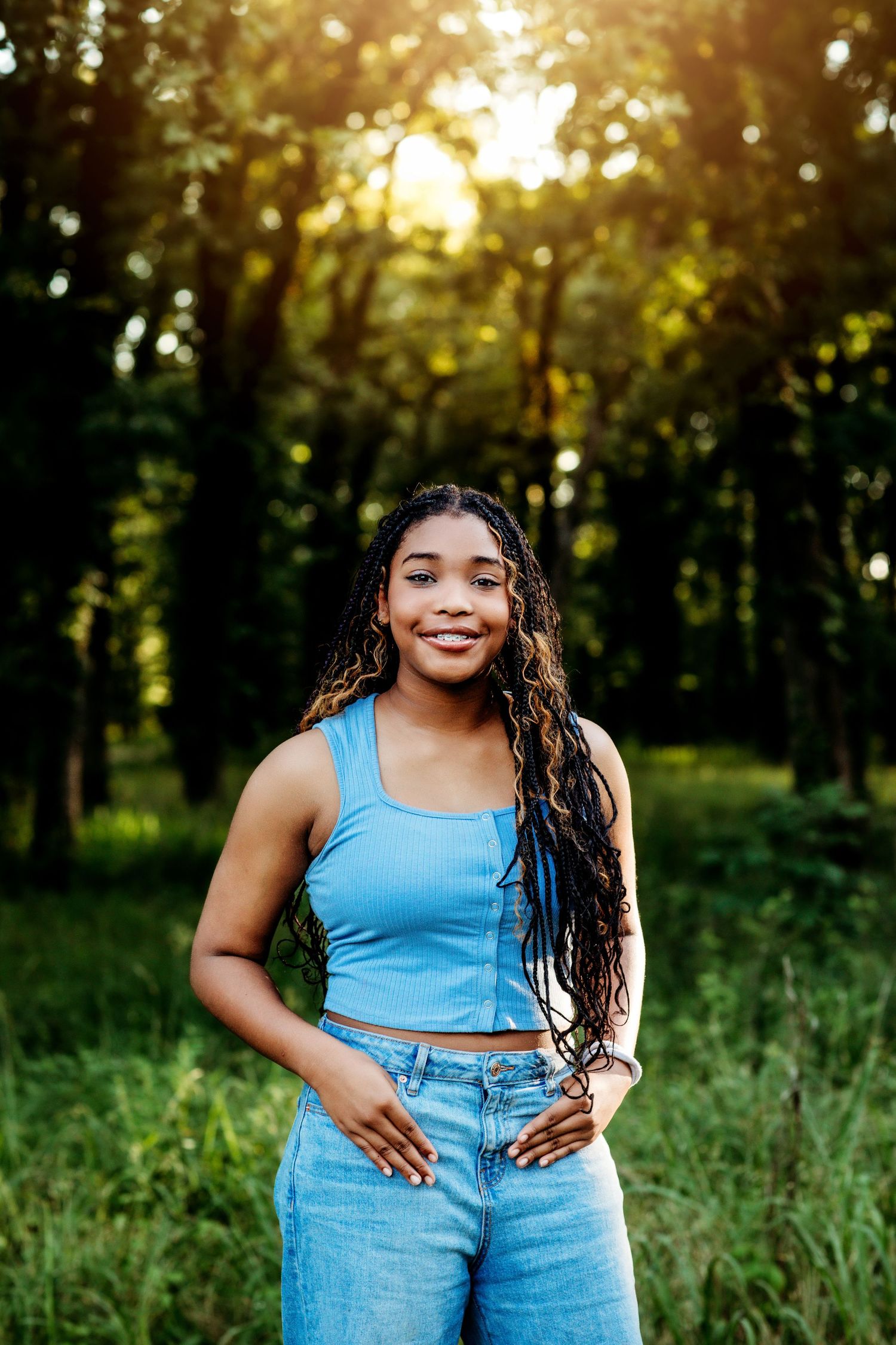 Young Black woman with braids smiles in a sunlit forest, wearing a blue crop top and jeans.