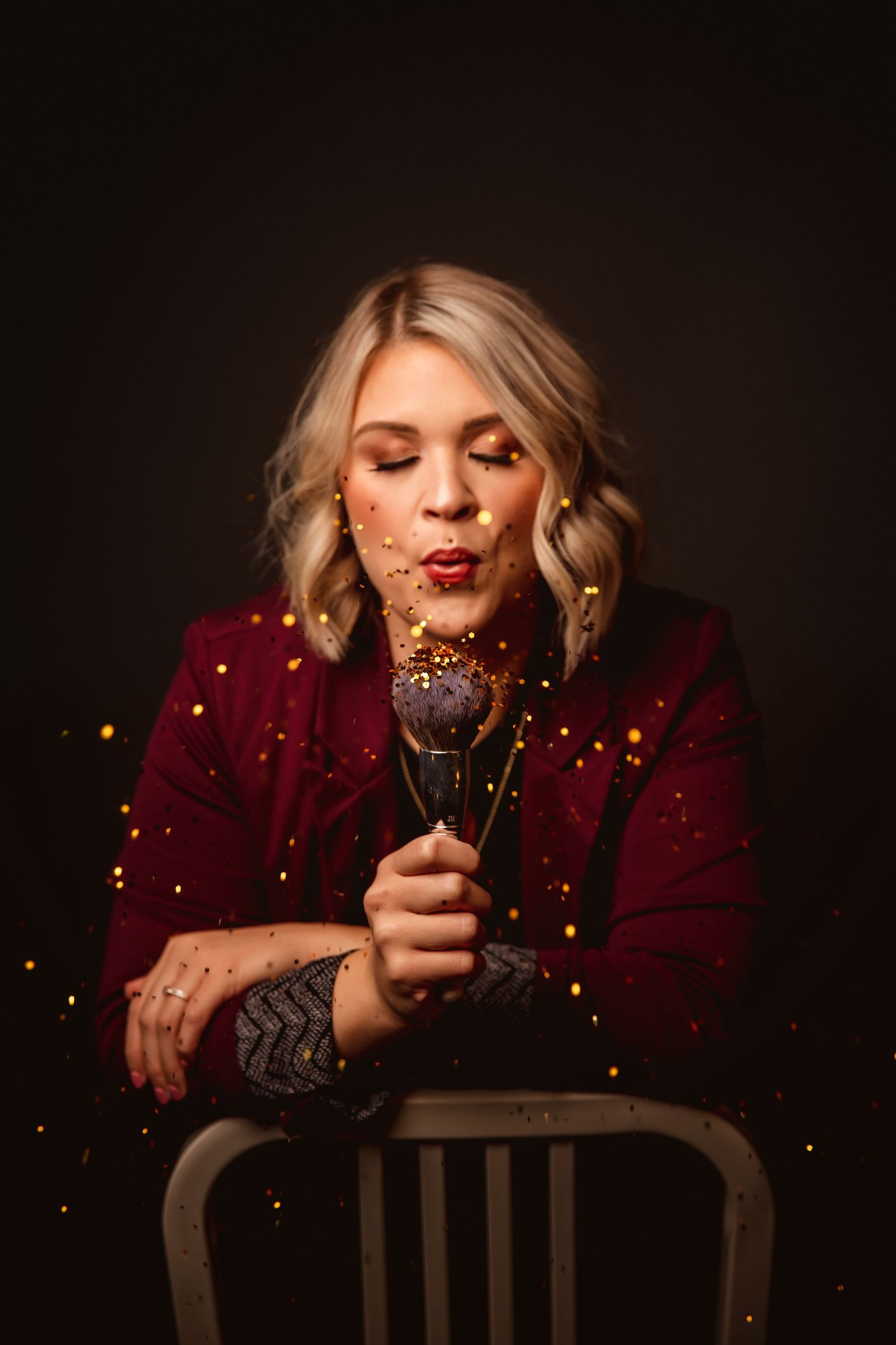 Blonde woman in burgundy jacket blowing glitter from a cake pop, studio portrait with dark background.