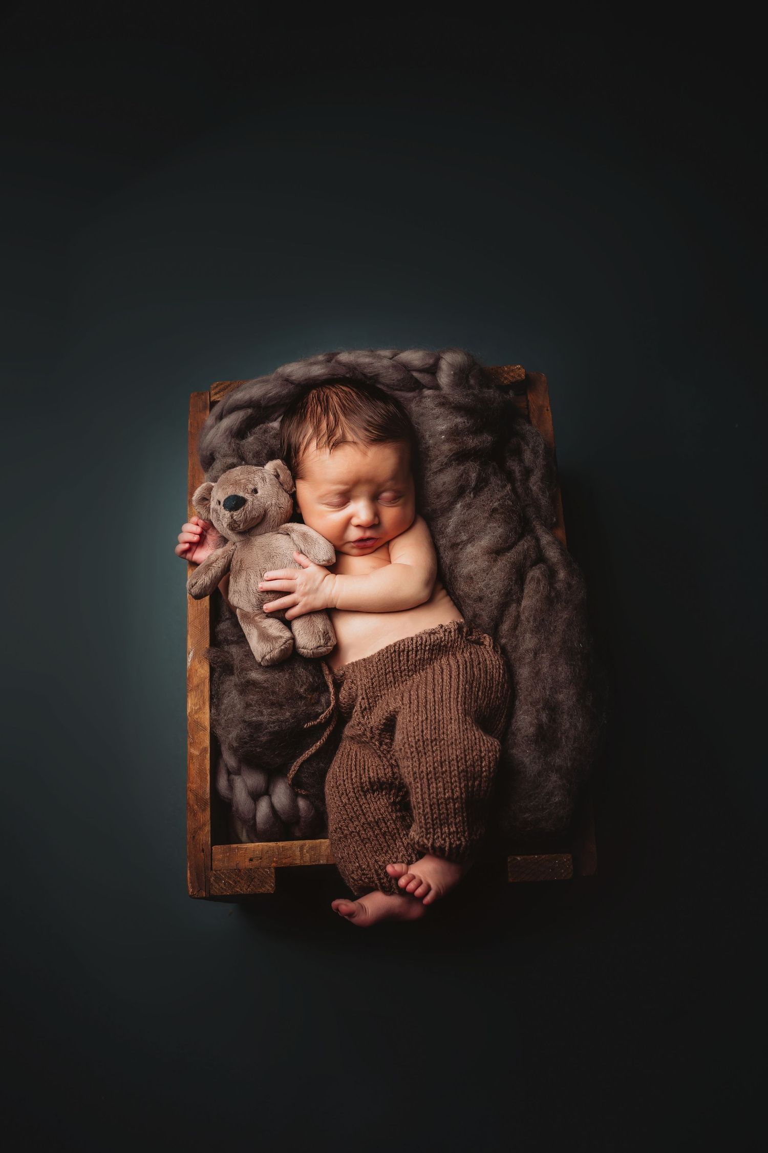 Newborn baby sleeping in wooden box with brown knit pants, hugging a teddy bear on a soft fur blanket.