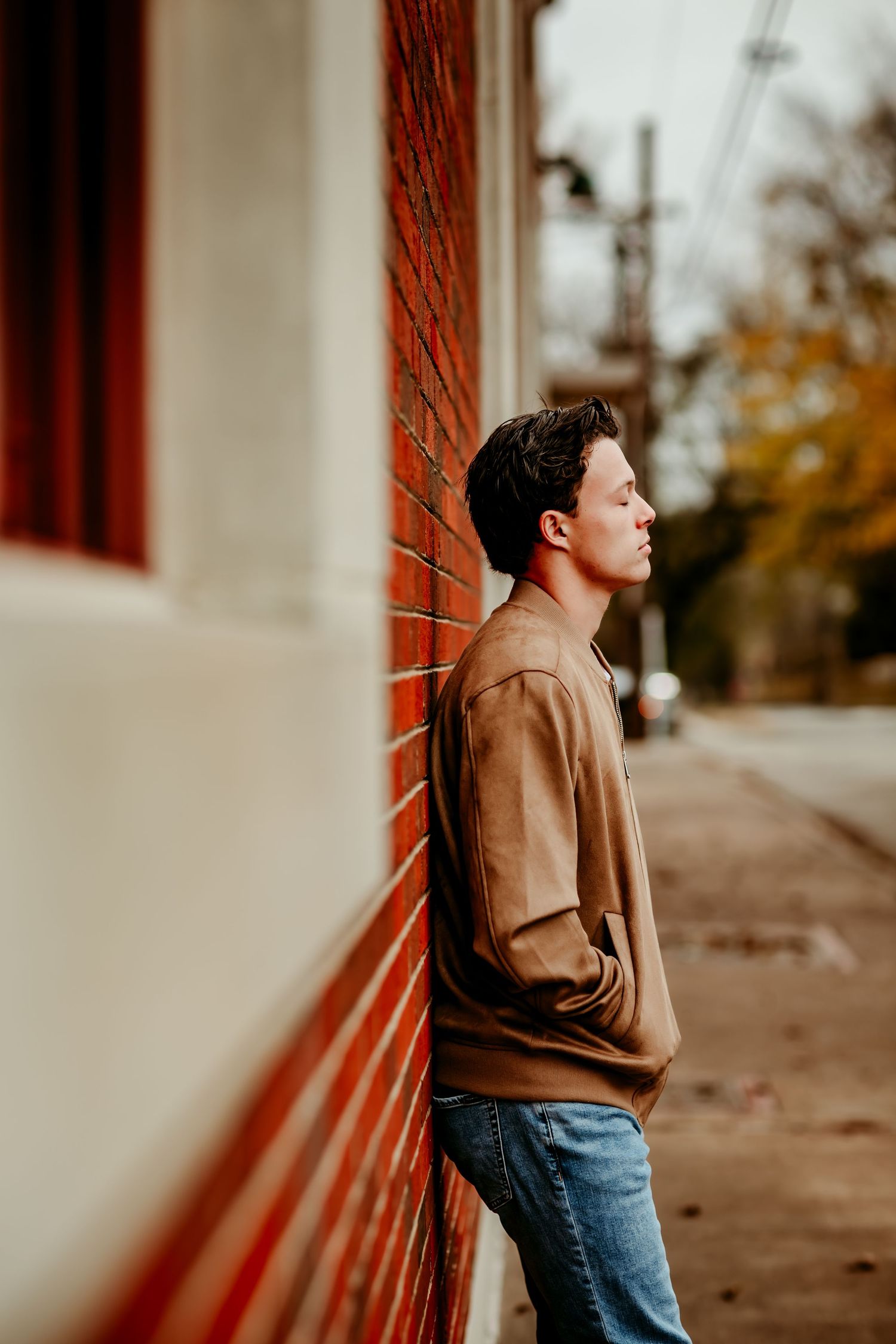 Young man in brown sweater and jeans leaning against red brick wall outdoors in autumn setting.