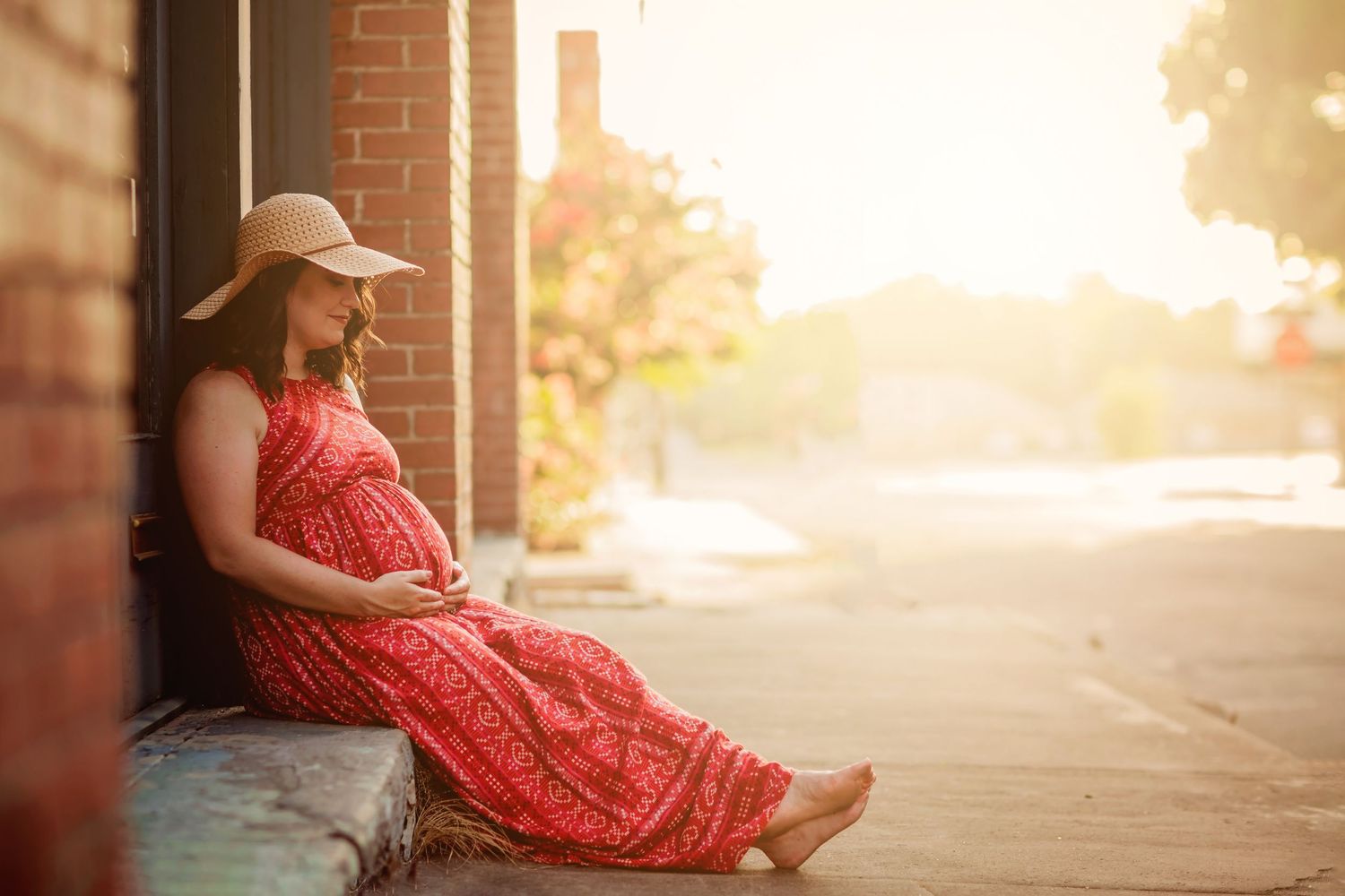 Pregnant woman in red floral dress and sun hat sits against brick wall in golden sunlight, cradling her baby bump.