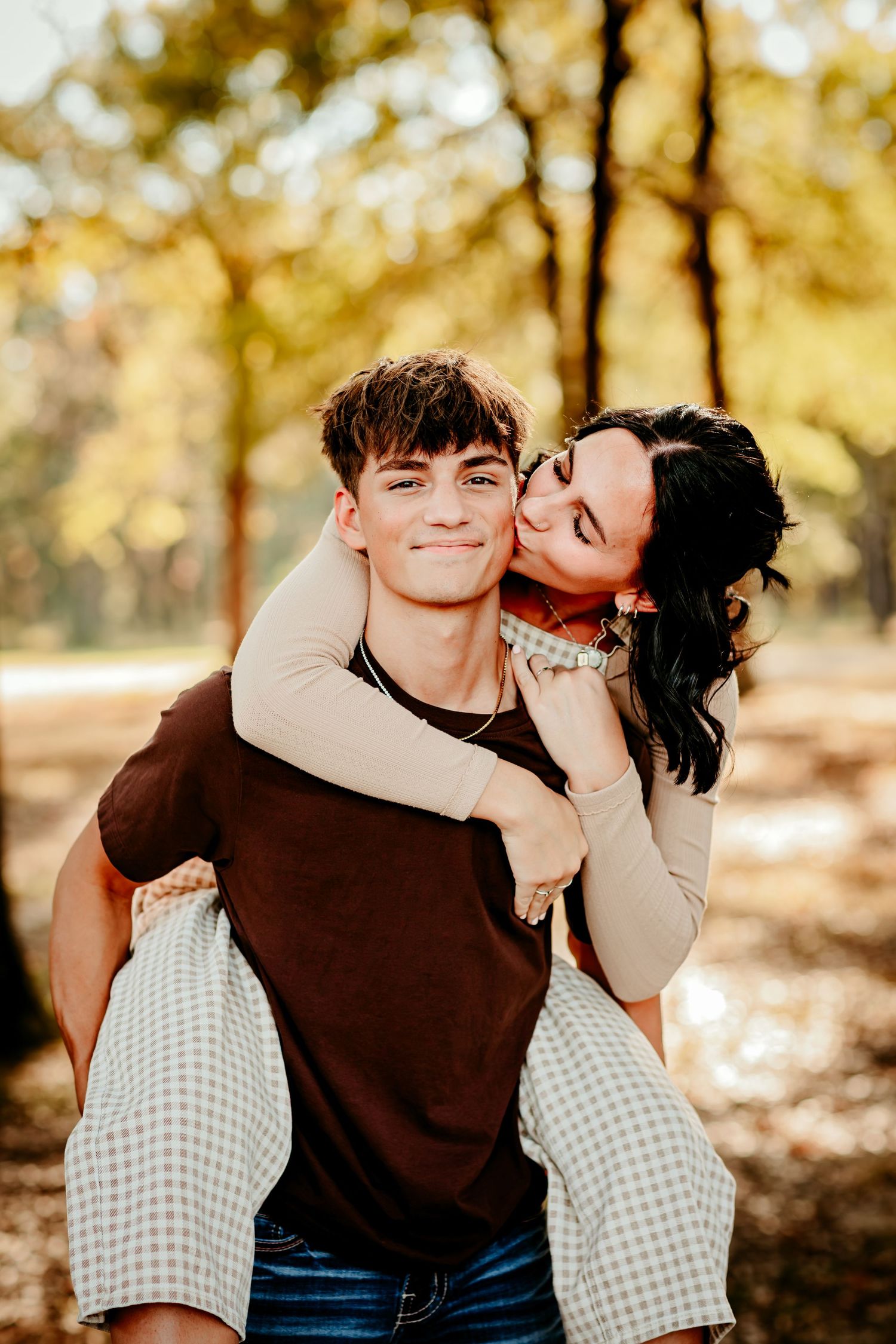 Young couple in autumn park, man giving woman piggyback ride while she kisses his cheek, smiling at camera.