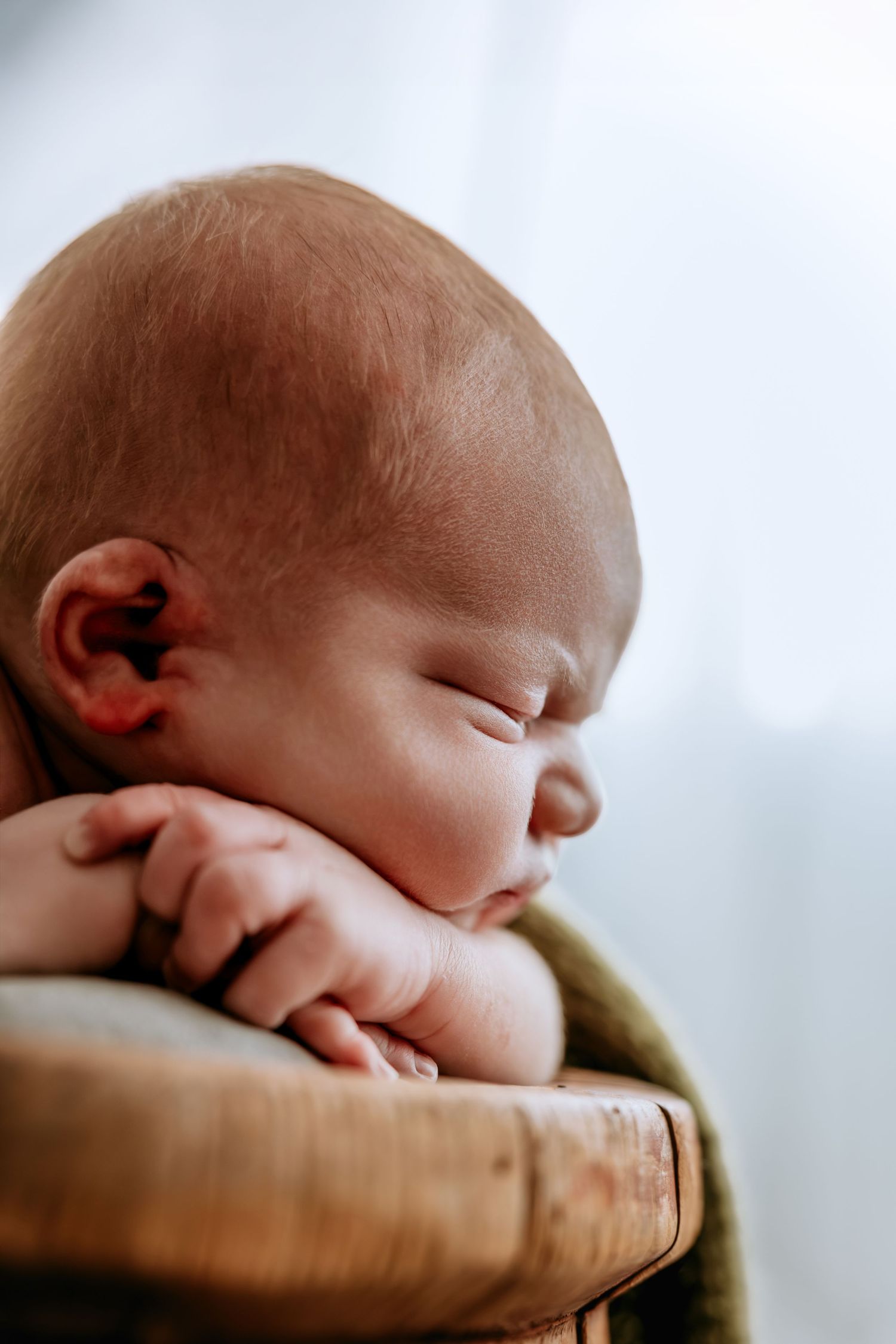 A peaceful newborn baby sleeping with tiny hands tucked under chin, resting in a wooden bowl with soft natural light.