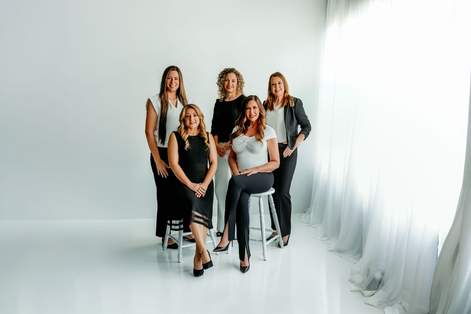 Five professional women pose together in a bright white studio, wearing elegant black and white outfits for a group portrait.