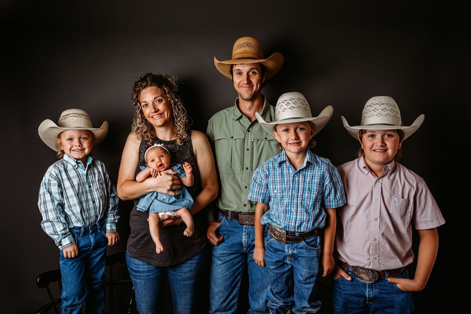 Family of six in cowboy hats posing for a studio portrait, parents holding newborn baby with three boys in western attire.