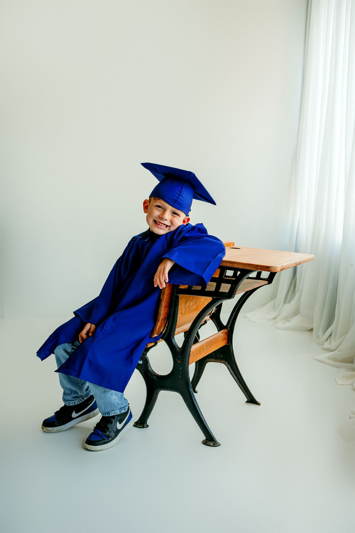 Smiling young boy in blue graduation cap and gown sitting at a vintage school desk.