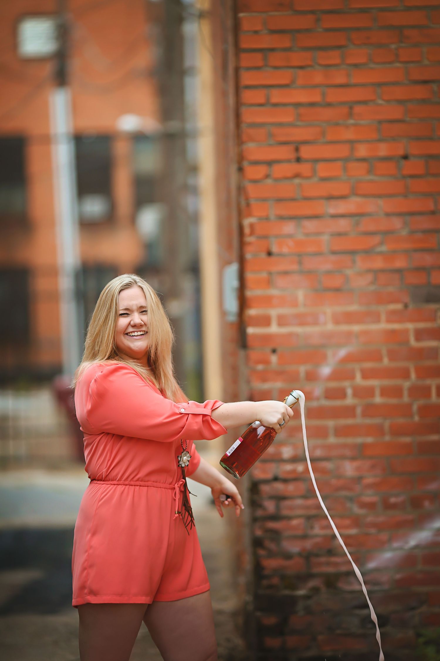 Smiling blonde woman in coral romper popping champagne bottle outdoors near red brick wall.