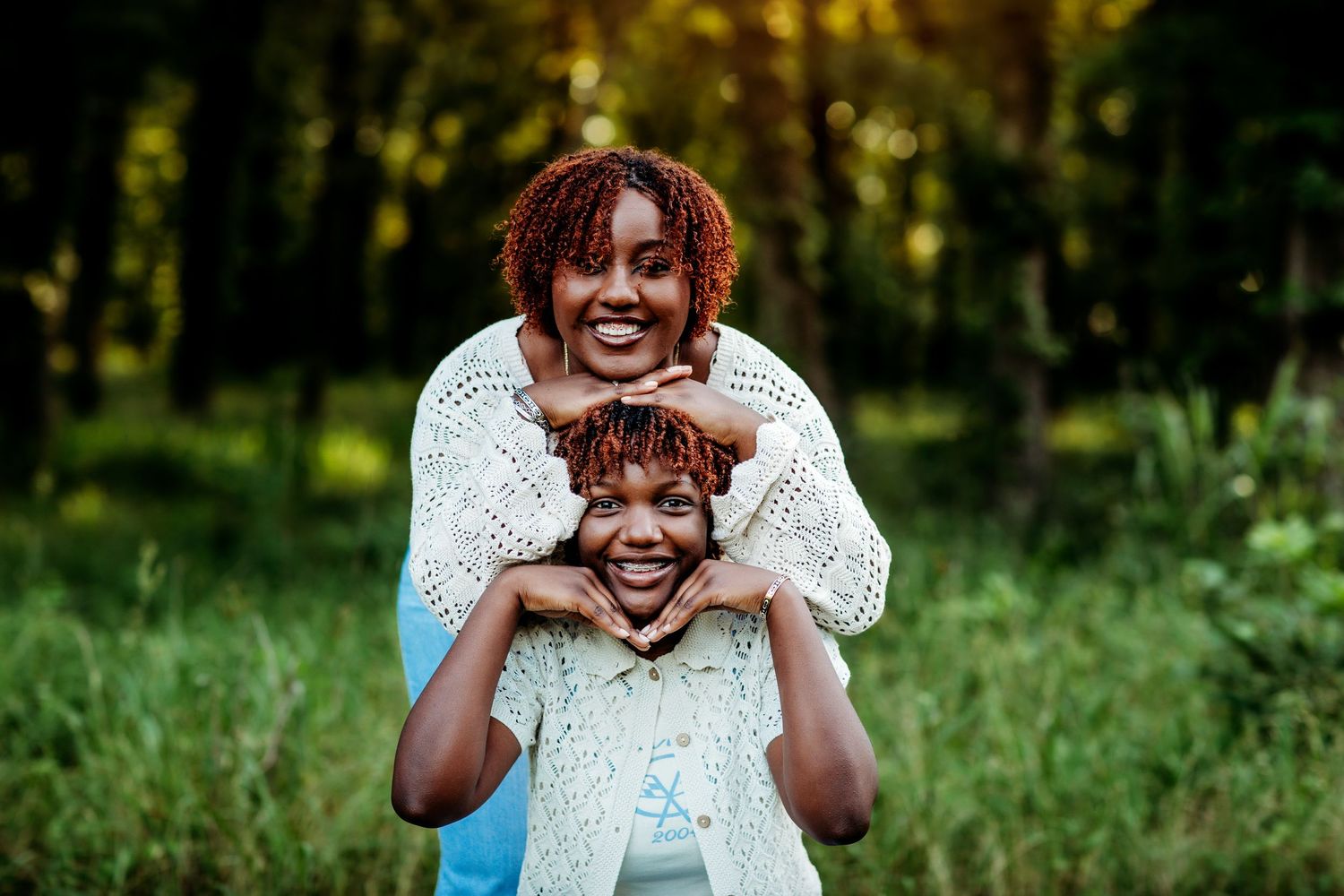 A joyful mother and daughter pose together outdoors, smiling in a lush green forest setting.