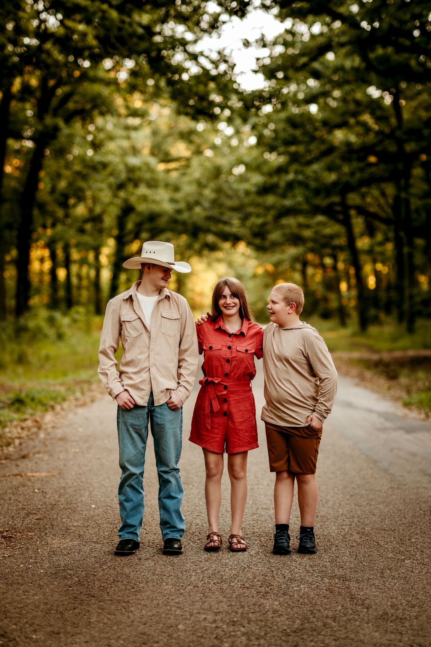 Three young people posing on a tree-lined dirt road, with one wearing a cowboy hat and a woman in a red dress.
