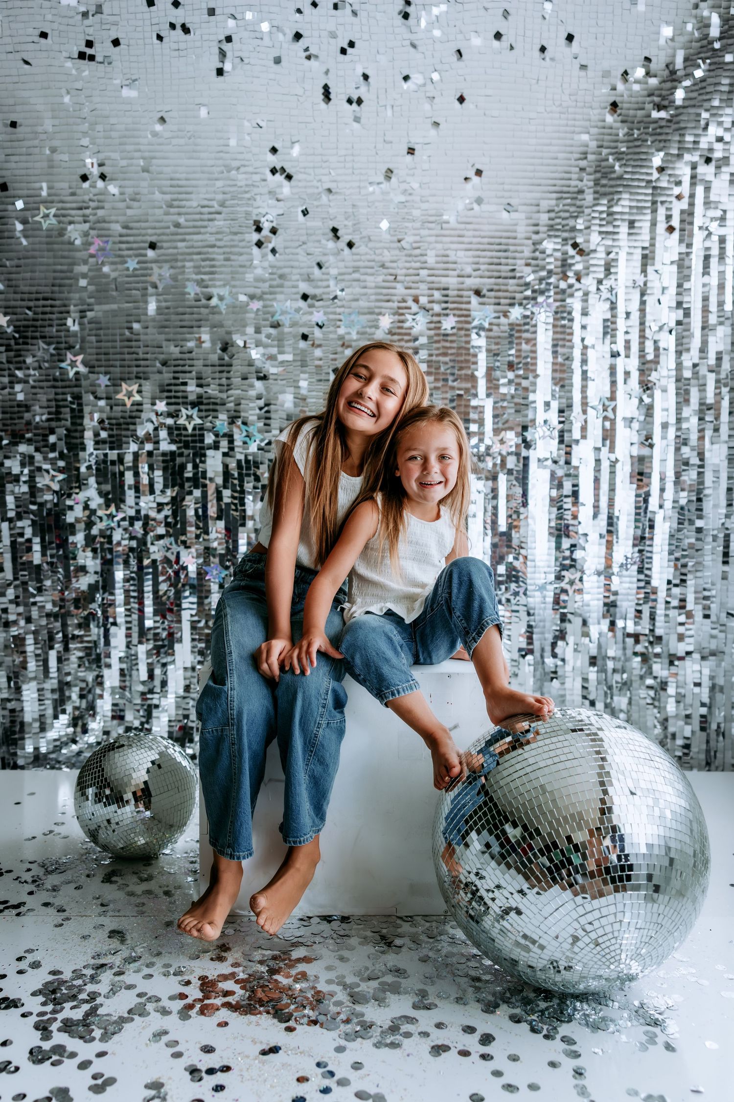 Two girls smiling and posing with disco balls and falling confetti against a shiny silver sequin wall.