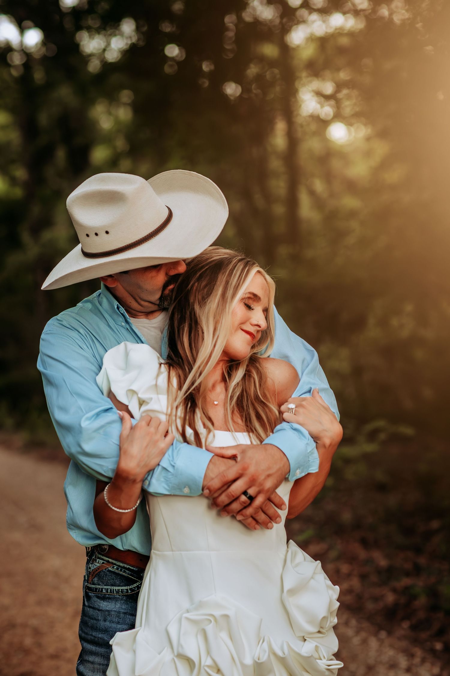 A romantic couple in western attire embraces during a sunset photoshoot in a wooded setting.