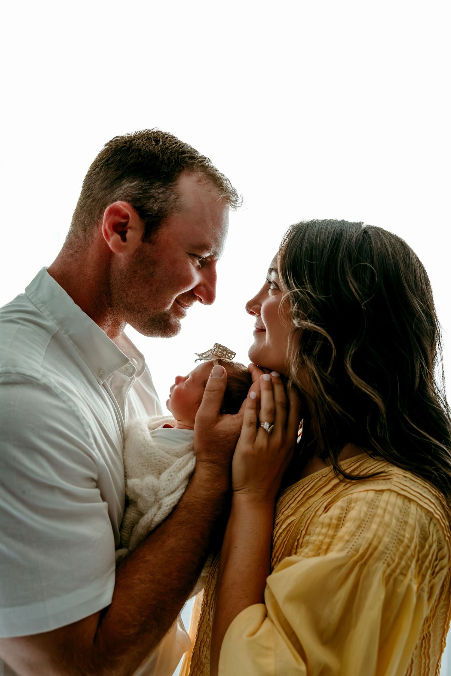 A couple tenderly gazes at each other while cradling their newborn baby in a bright, softly lit portrait.
