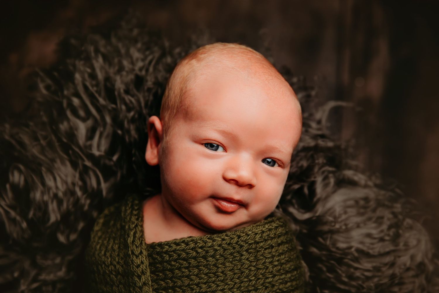 Newborn baby wrapped in olive green knit blanket against dark textured background.