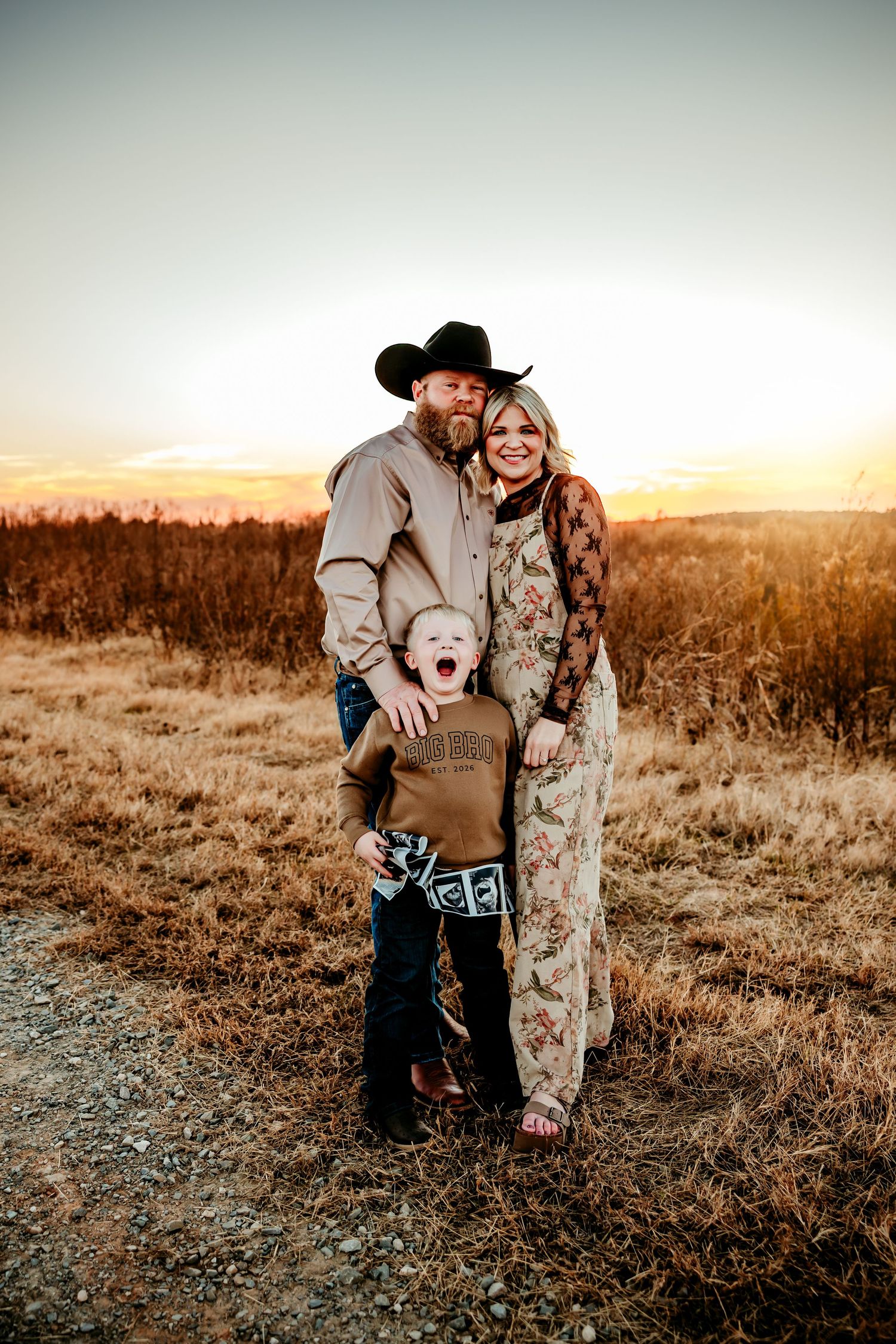 Family portrait at golden hour with man in cowboy hat, woman in floral jumpsuit, and young boy posing on a rural dirt path.