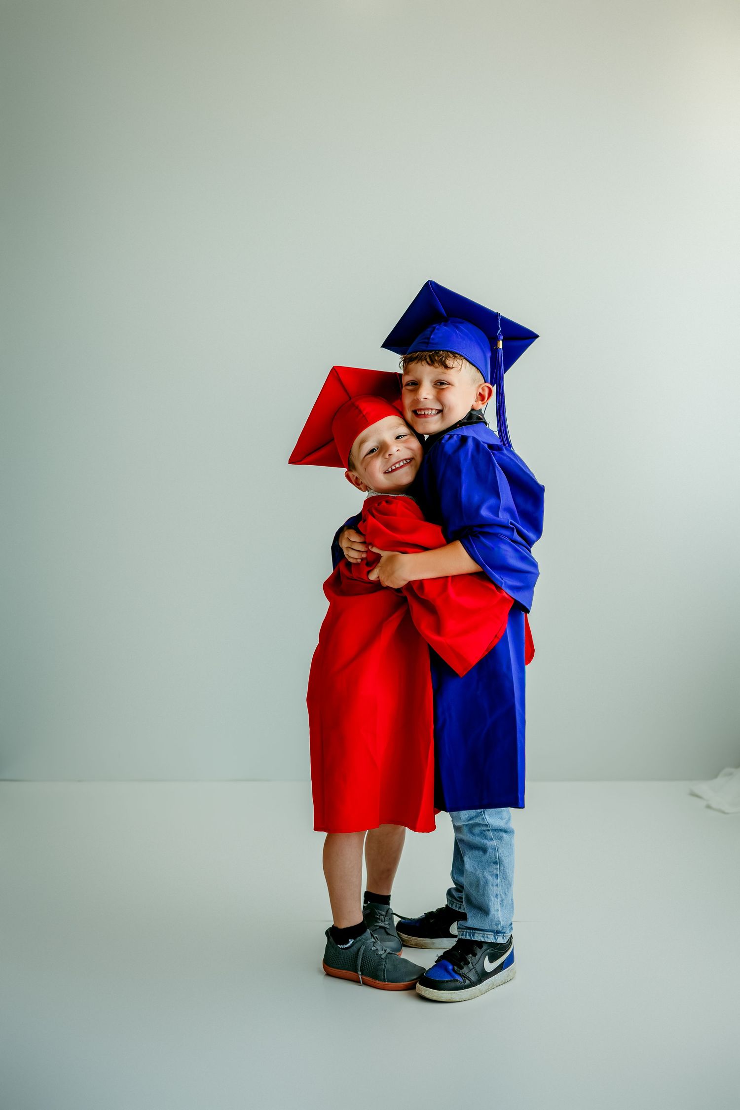Two young boys in red and blue graduation caps and gowns hugging and smiling against a white background.