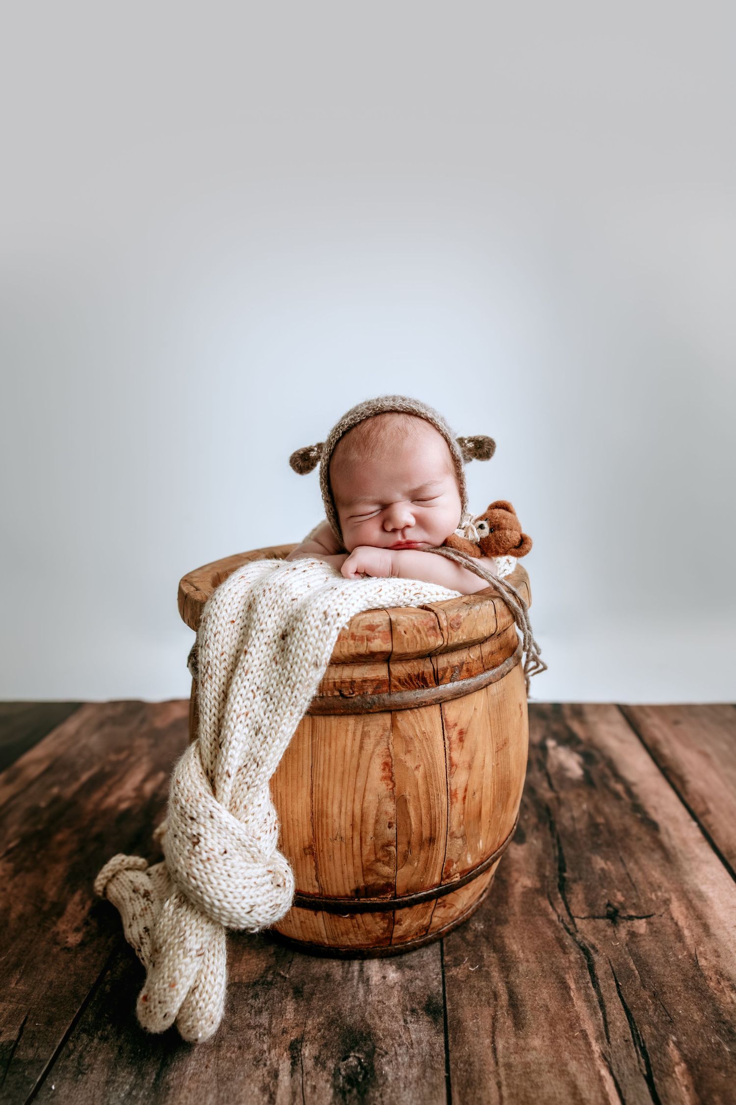 Sleeping newborn baby in a wooden barrel bucket wearing a knit bear bonnet hat on a rustic wood floor.