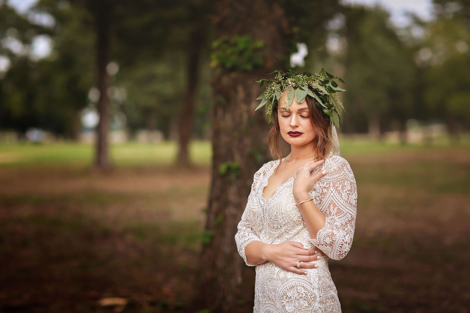 Bride in lace gown and green leaf crown poses gracefully outdoors among trees in a serene garden setting.