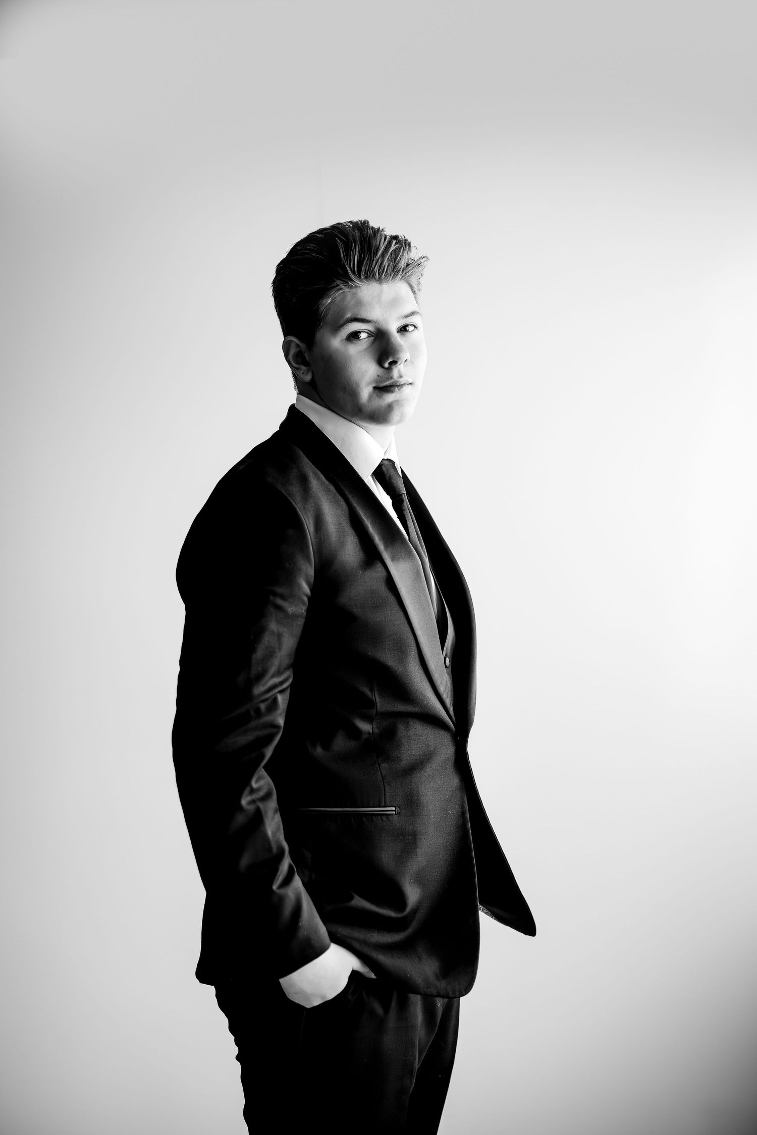 Black and white portrait of a young man in a dark suit and tie, hands in pockets, posing confidently against a light background.