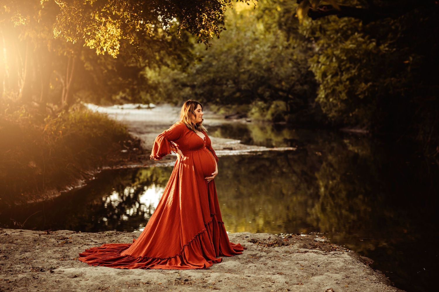 Pregnant woman in flowing red gown standing by sunlit river surrounded by lush green trees during golden hour.