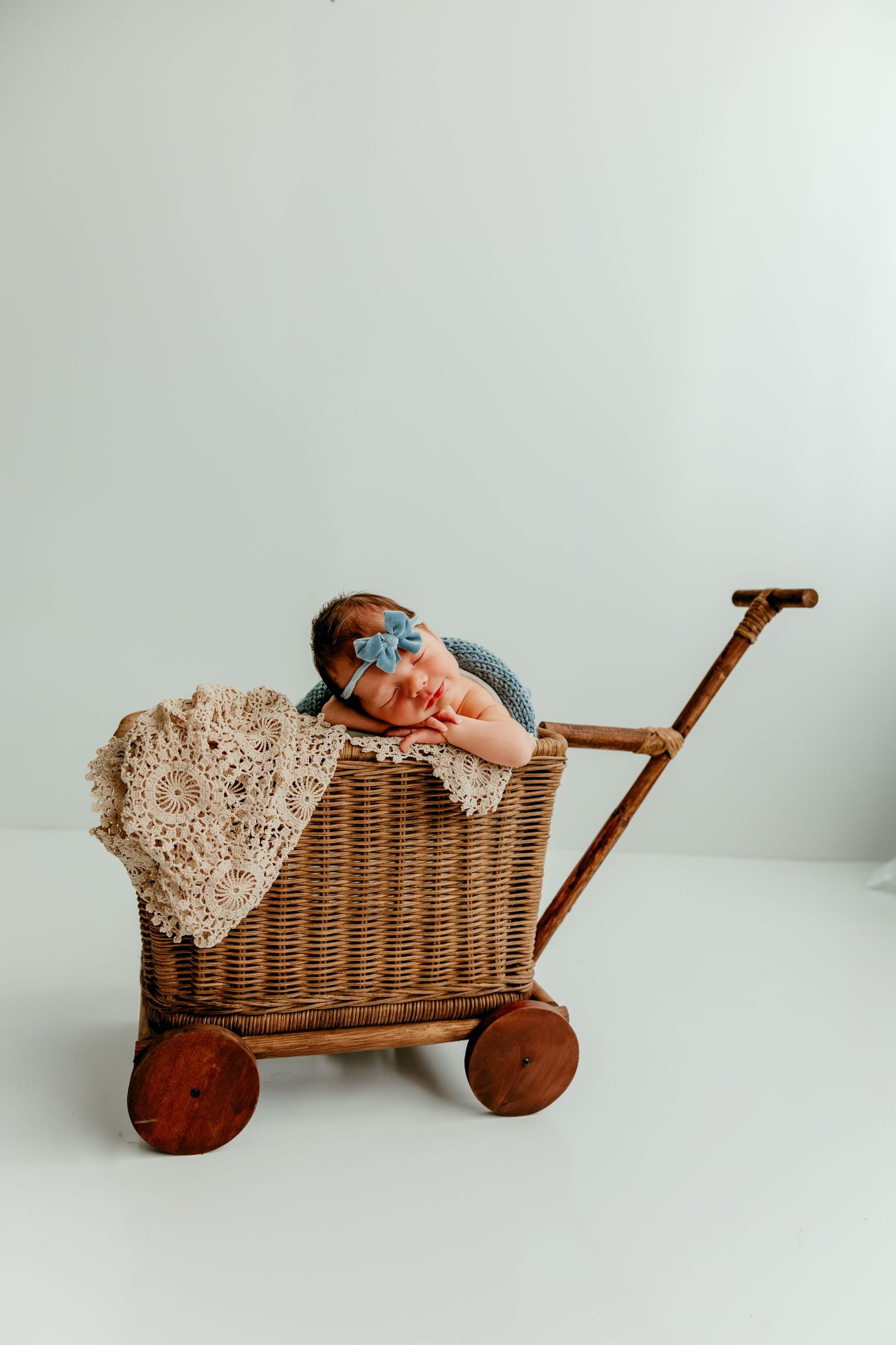 Newborn baby sleeping in a vintage wicker cart with lace blanket and blue bow headband on white background.