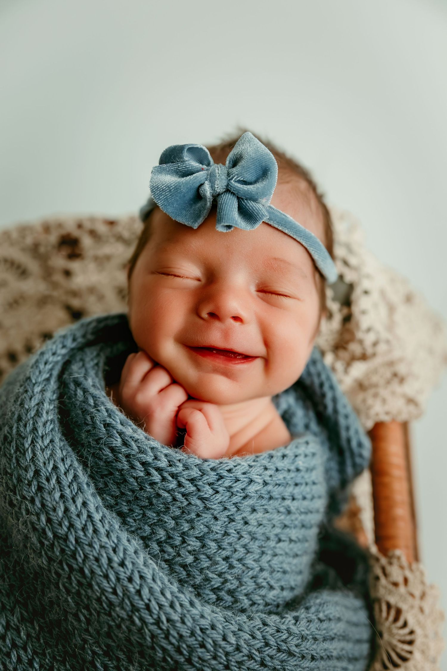 Smiling newborn baby wrapped in a blue knit blanket with a matching bow headband, sitting in a wicker basket.