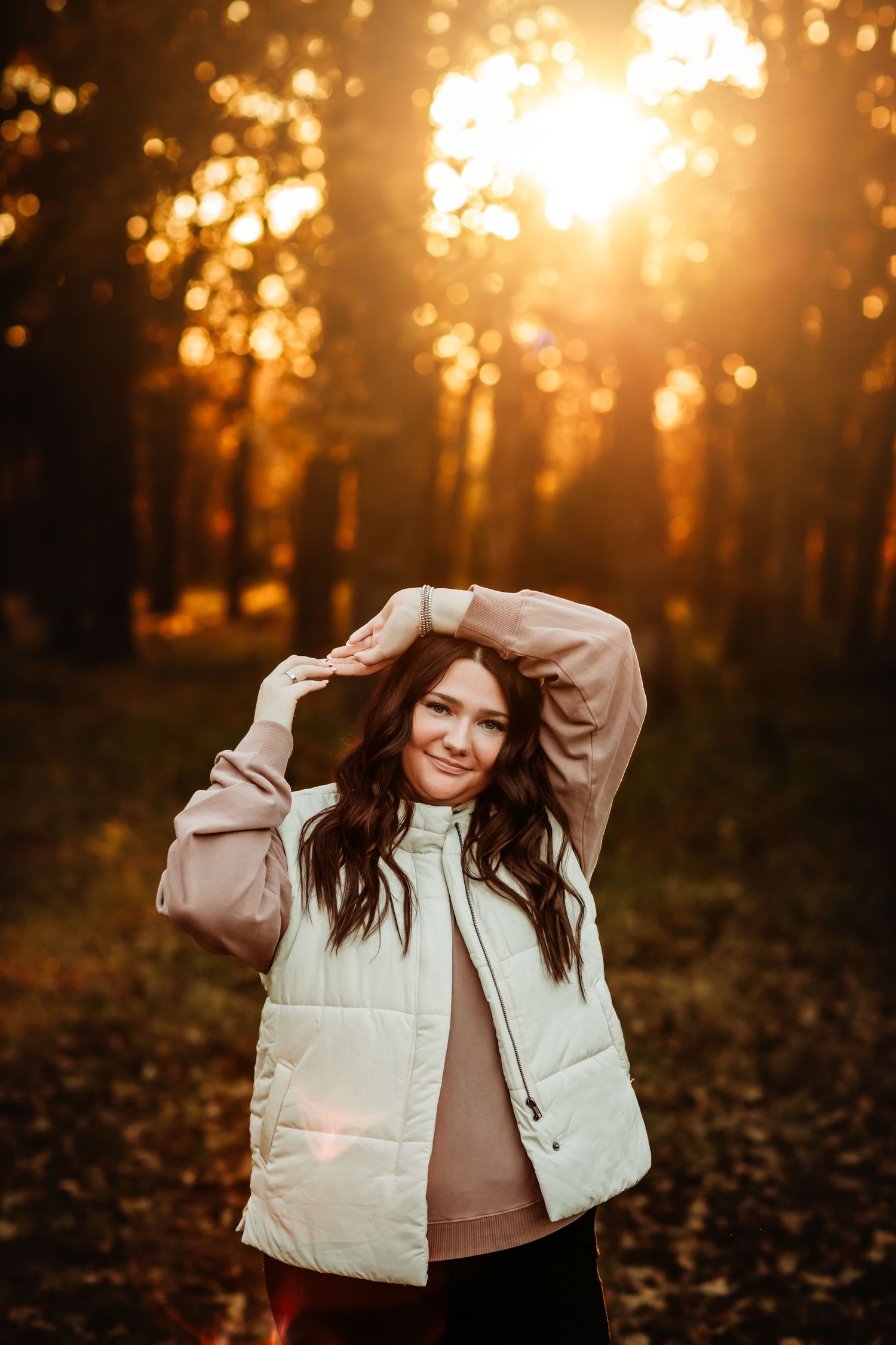 Woman in white vest smiling in autumn forest with golden sunset light filtering through trees.