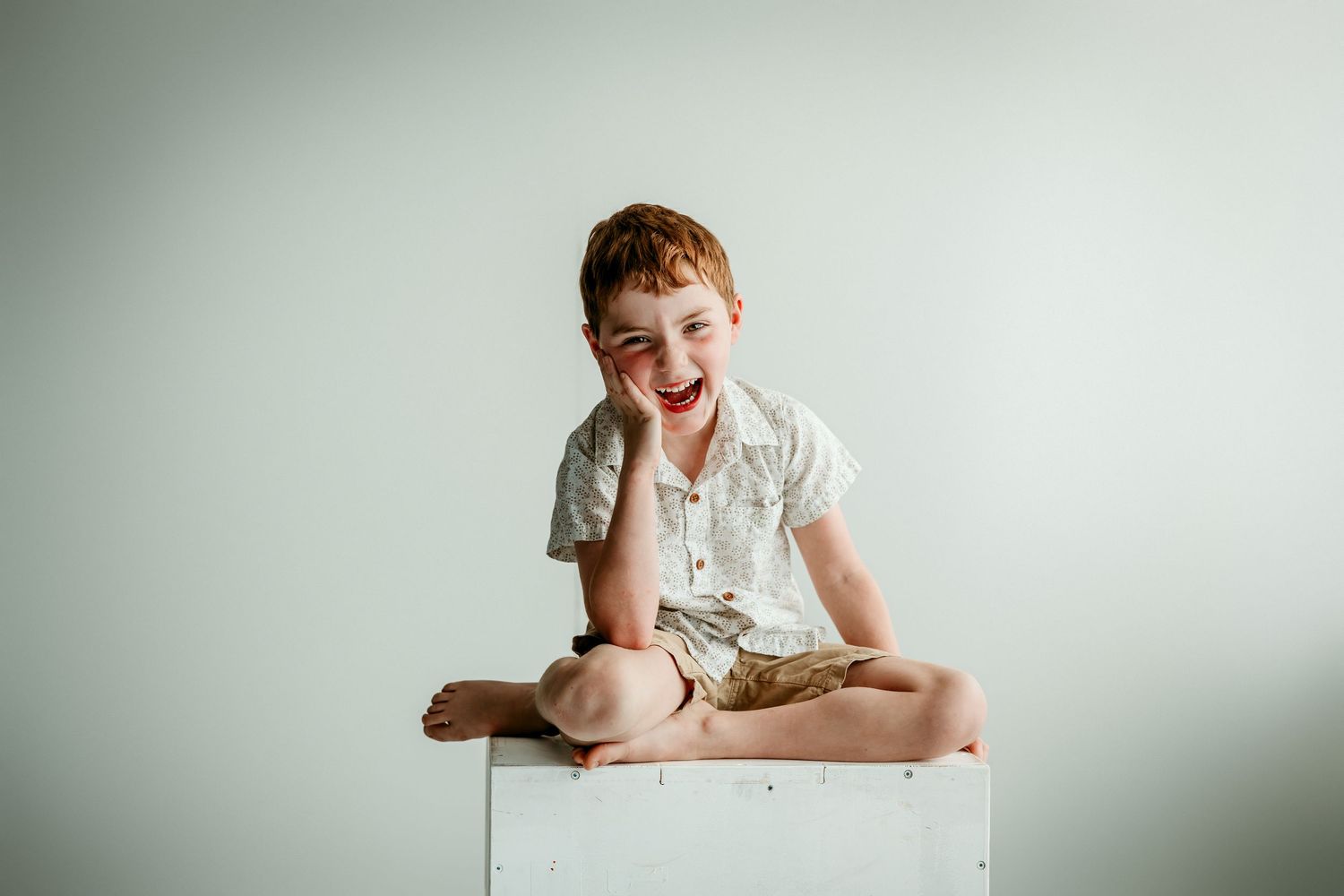 A laughing red-haired boy sits cross-legged on a white box against a light gray background.