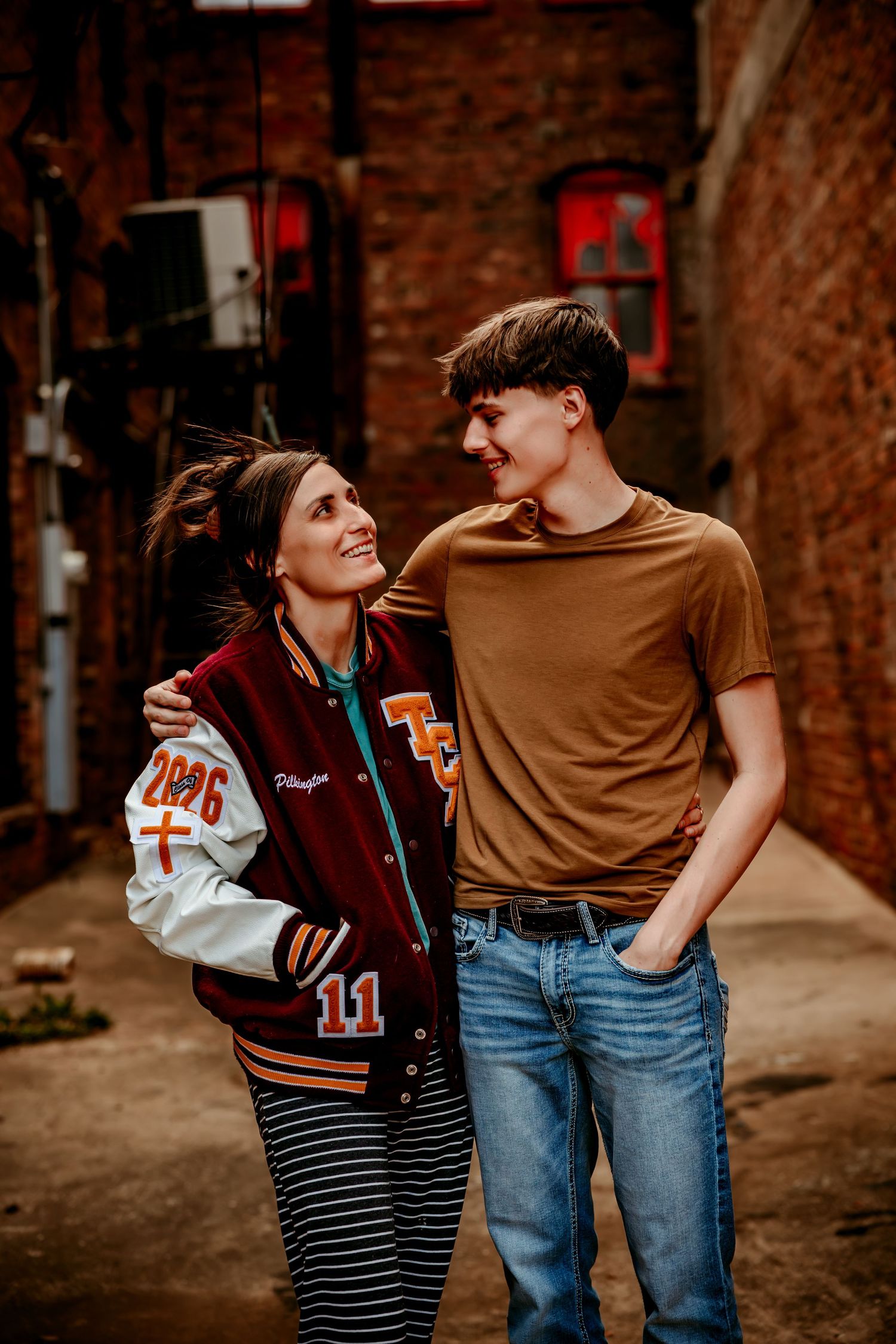 A young couple smiles at each other in a brick alley, she wears a varsity jacket and striped skirt, he wears a tan tee and jeans.